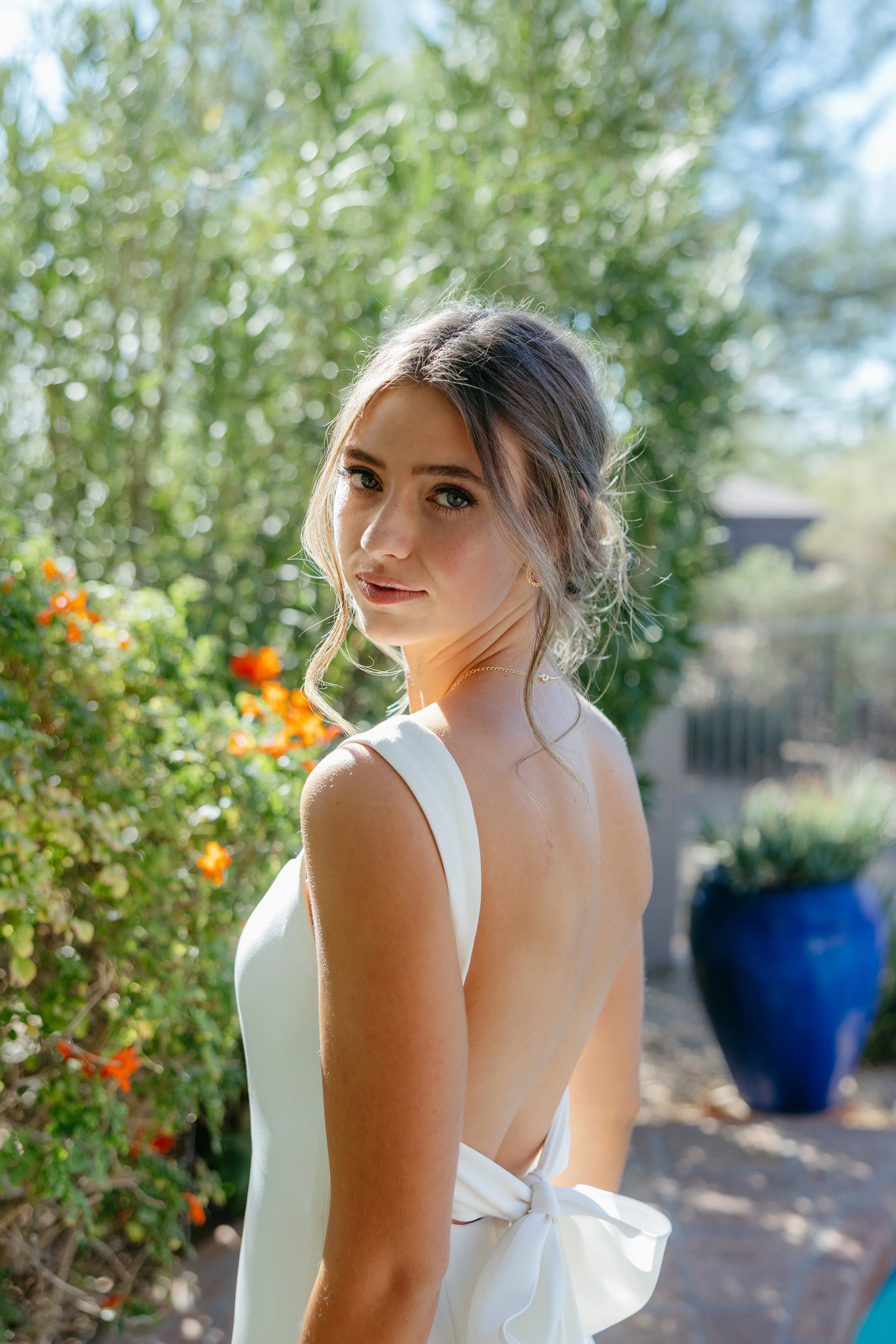A young woman with wavy brown hair and blue eyes, wearing a white sleeveless dress with a back bow, standing outdoors in a garden with sunlight filtering through green foliage and orange flowers.