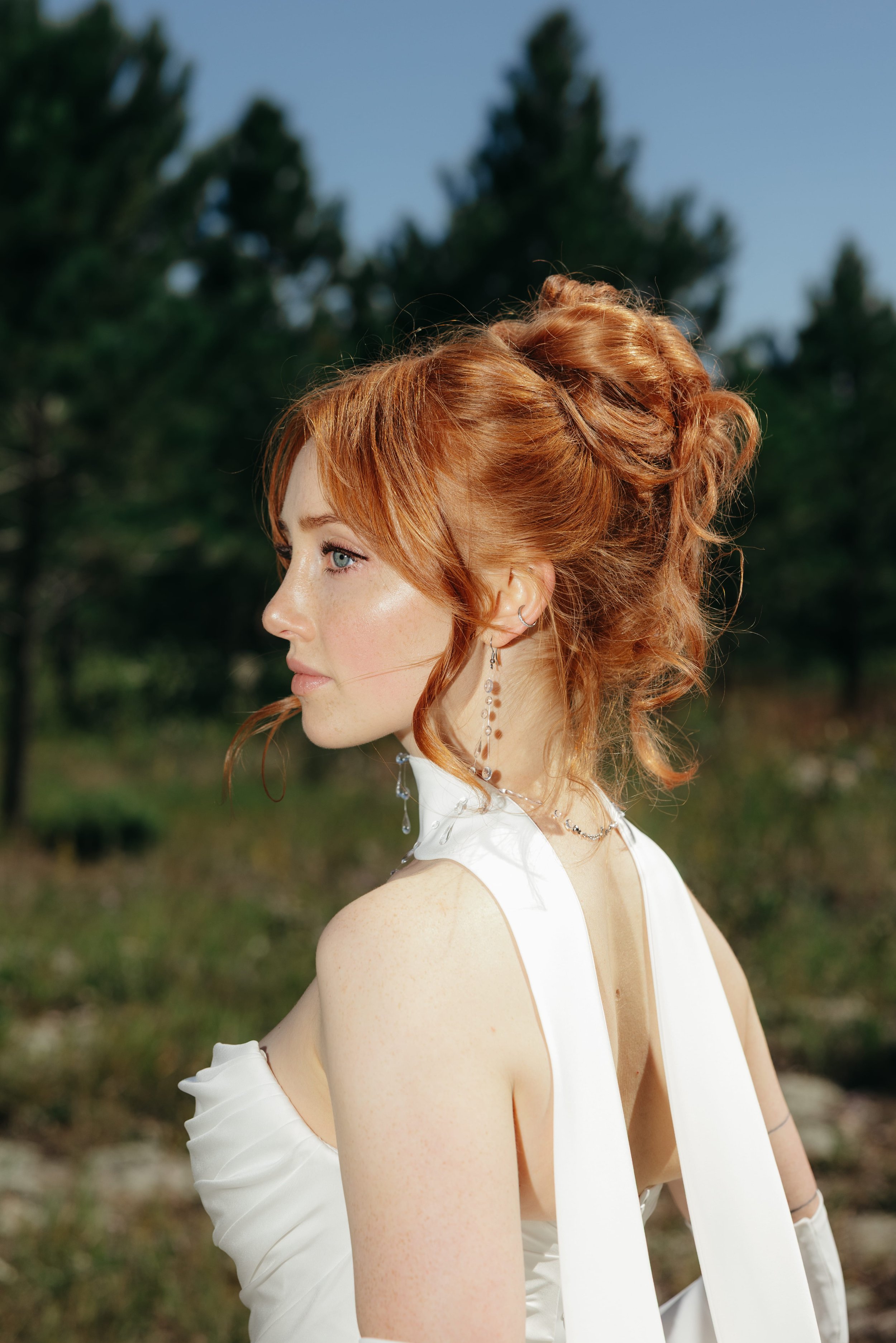 Profile of a young woman with curly red hair, wearing a white dress and dangling earrings, outdoors with a blurred green and blue background.