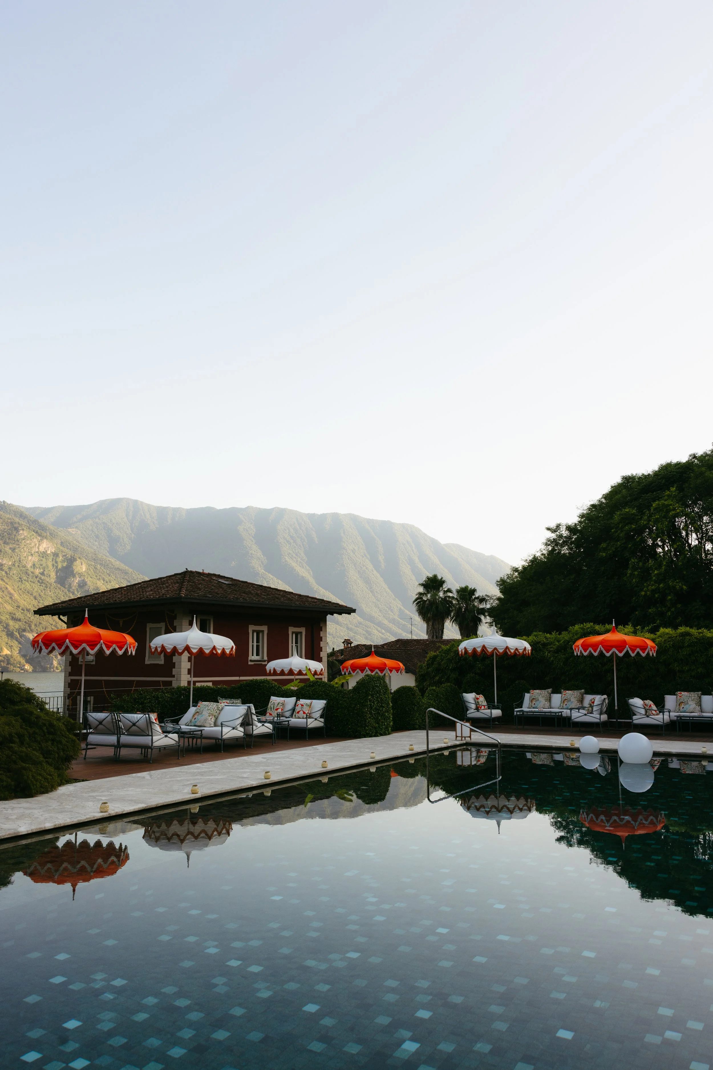 A serene outdoor pool area with red and white umbrellas, lounge chairs with cushions, surrounded by lush greenery and mountains in the background.