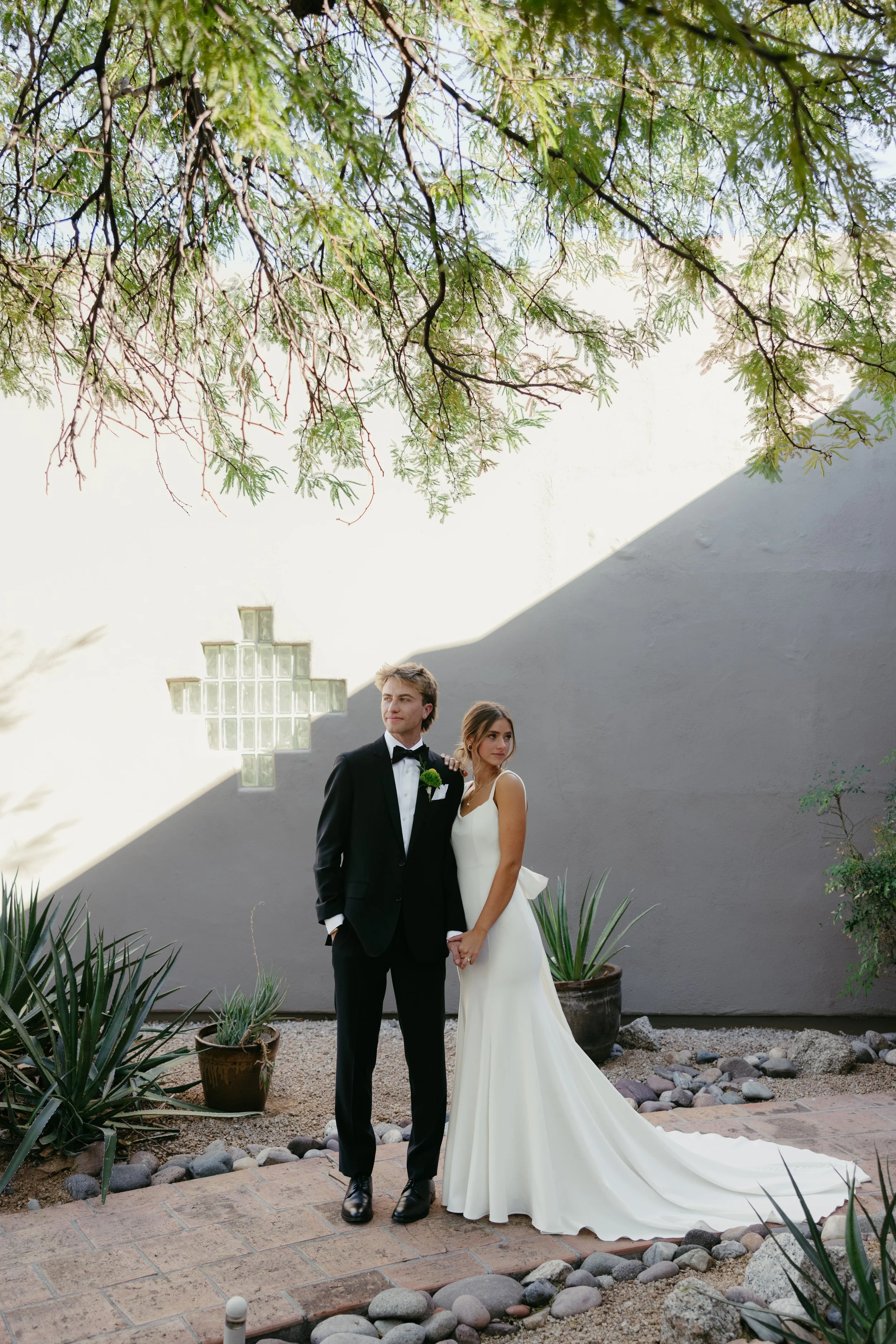 Bride and groom standing hand in hand outdoors, bride in a white wedding gown and groom in a black tuxedo, with plants and a partial wall with a glass block window in the background.