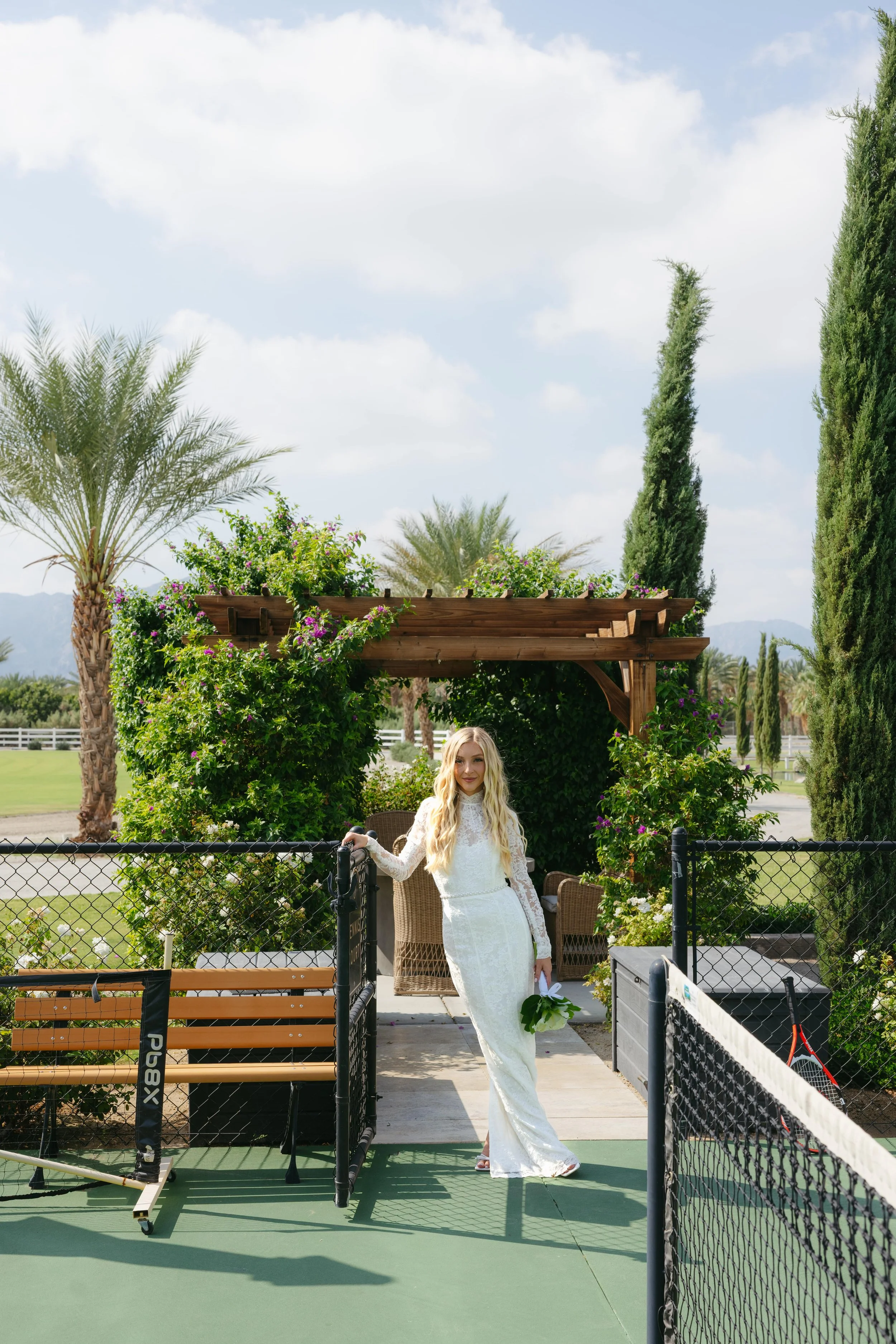 A woman in a white lace wedding dress holds a bouquet and stands by a tennis net outdoors on a sunny day, with palm and cypress trees and a wooden arbor decorated with greenery and purple flowers in the background.