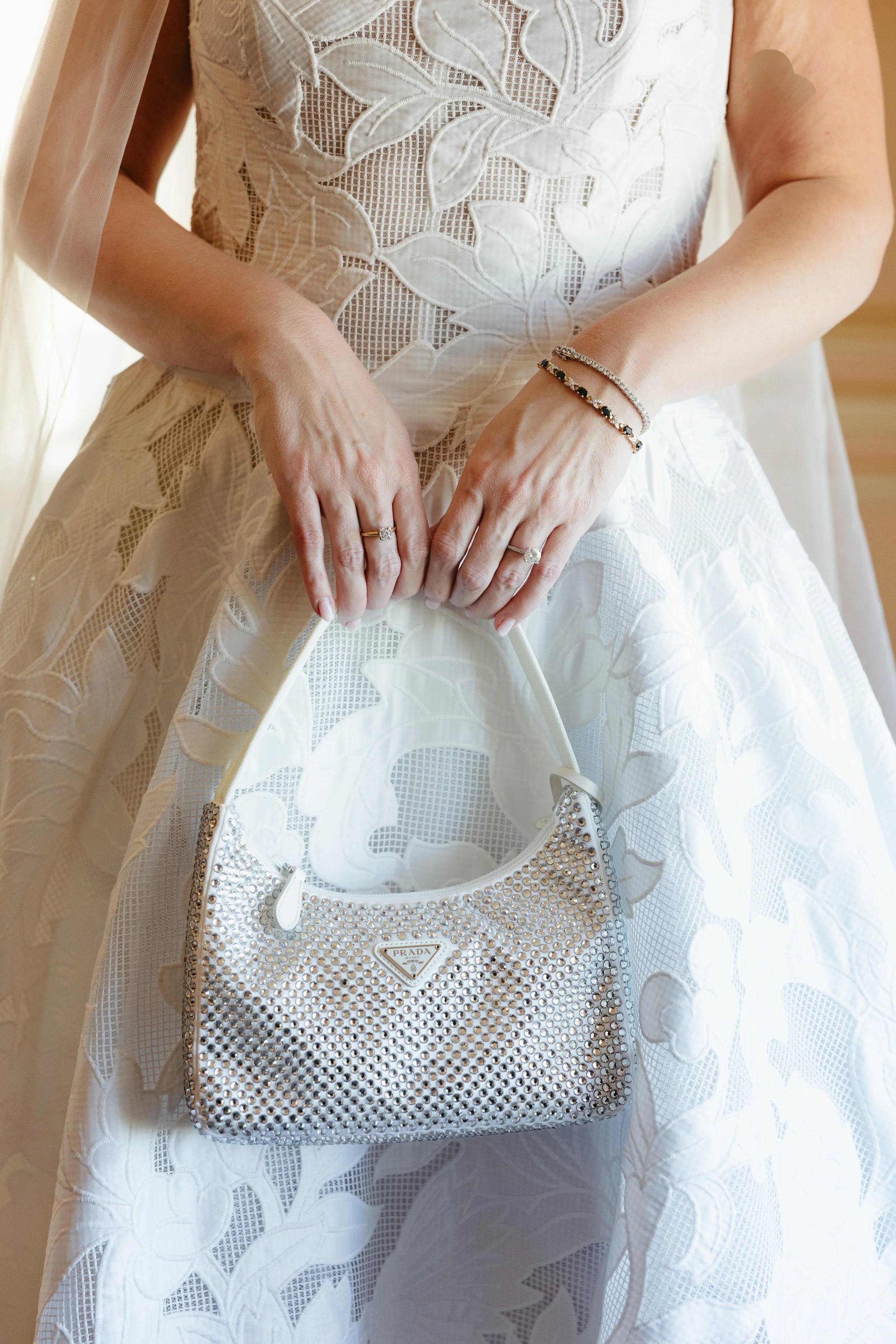 Close-up of a bride in a lace wedding dress holding a shiny Prada handbag, showing wedding rings and bracelets.