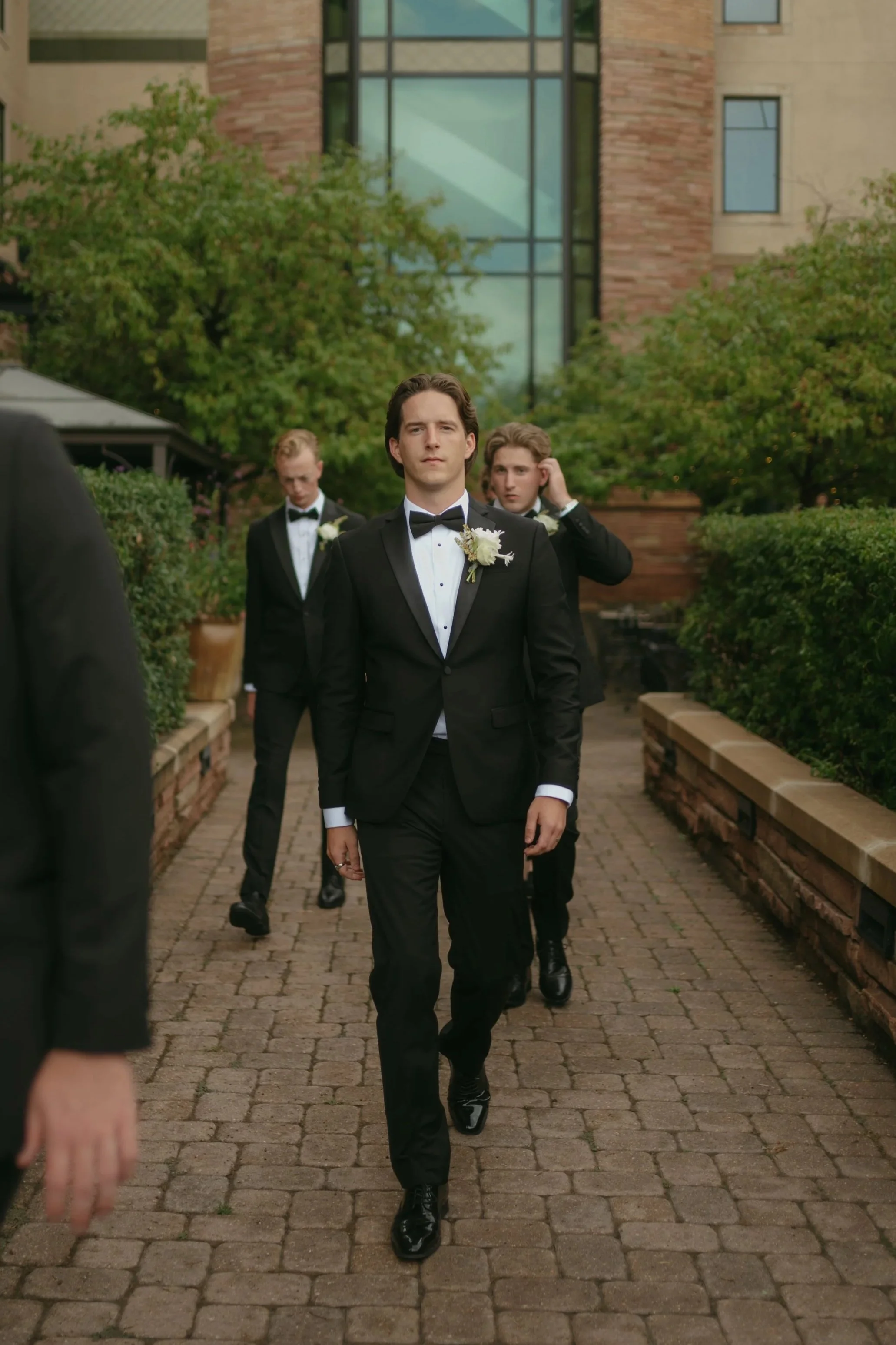 A group of men dressed in black tuxedos walking along a brick pathway outdoors during a formal event, with one man leading and the others following behind him.