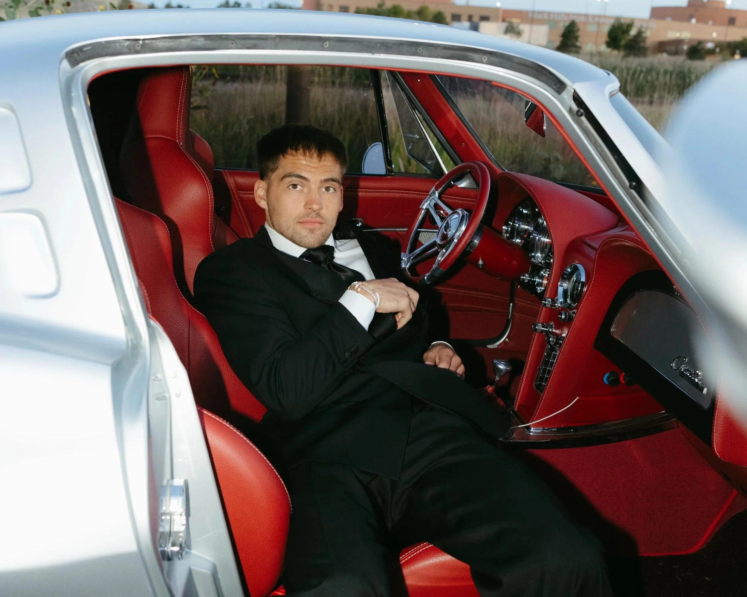 A man in a tuxedo sitting inside a classic vintage car with red interior, looking towards the camera.