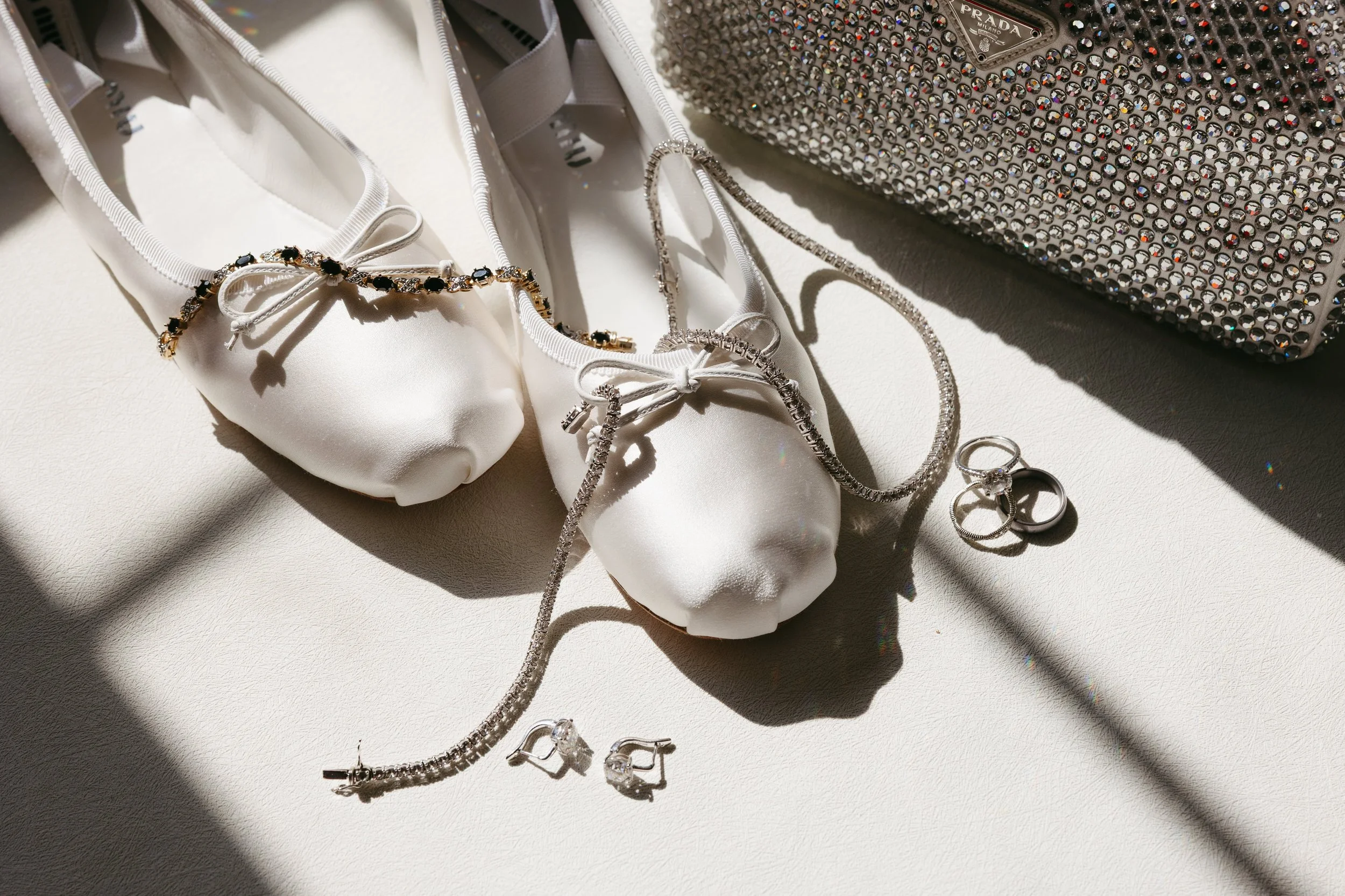 Silver jewelry including necklaces, earrings, and rings, along with white satin ballet flats with bows, arranged on a white surface with natural light and shadows, and diamond-studded accessory bag in the background.