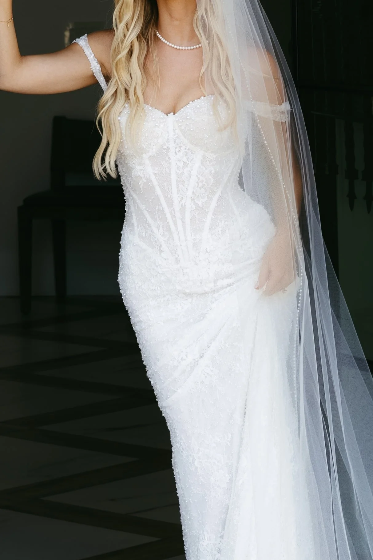Close-up of a bride in a white wedding gown with detailed lace and beading, holding up the skirt with one hand, wearing a pearl necklace and a veil, with a dark background.
