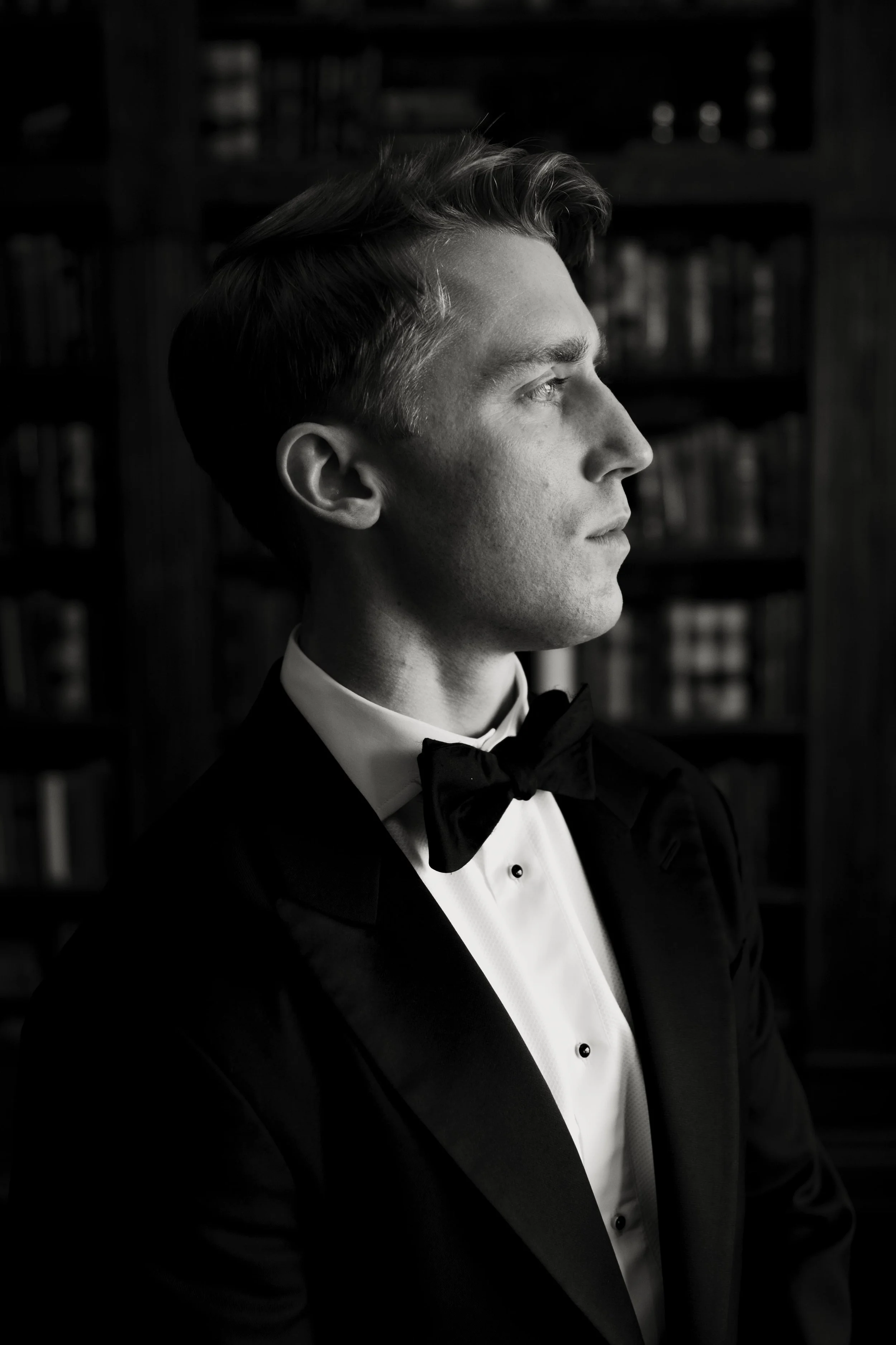 A black-and-white photo of a young man in a tuxedo and bow tie looking to the right, with a serious expression, in a room with a background of bookshelves.