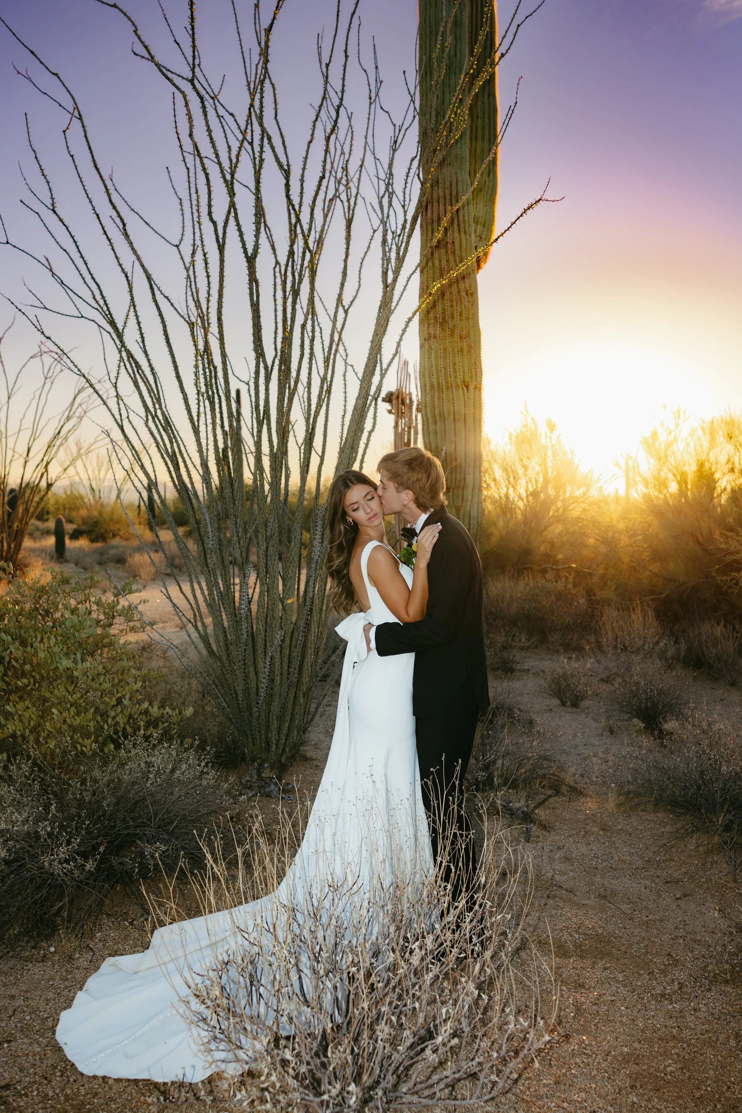 A bride and groom embracing in a desert landscape at sunset, with cacti and dry brush around them.