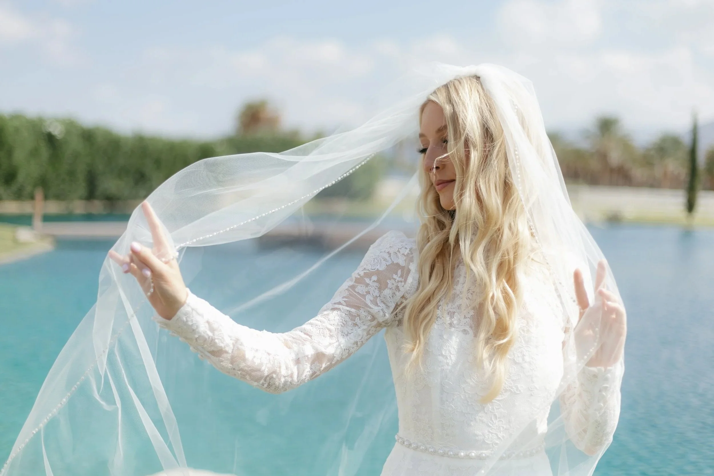 A woman with long blonde hair, wearing a white lace wedding dress, holding a veil near her face outdoors near a body of water, with trees and blue sky in the background.