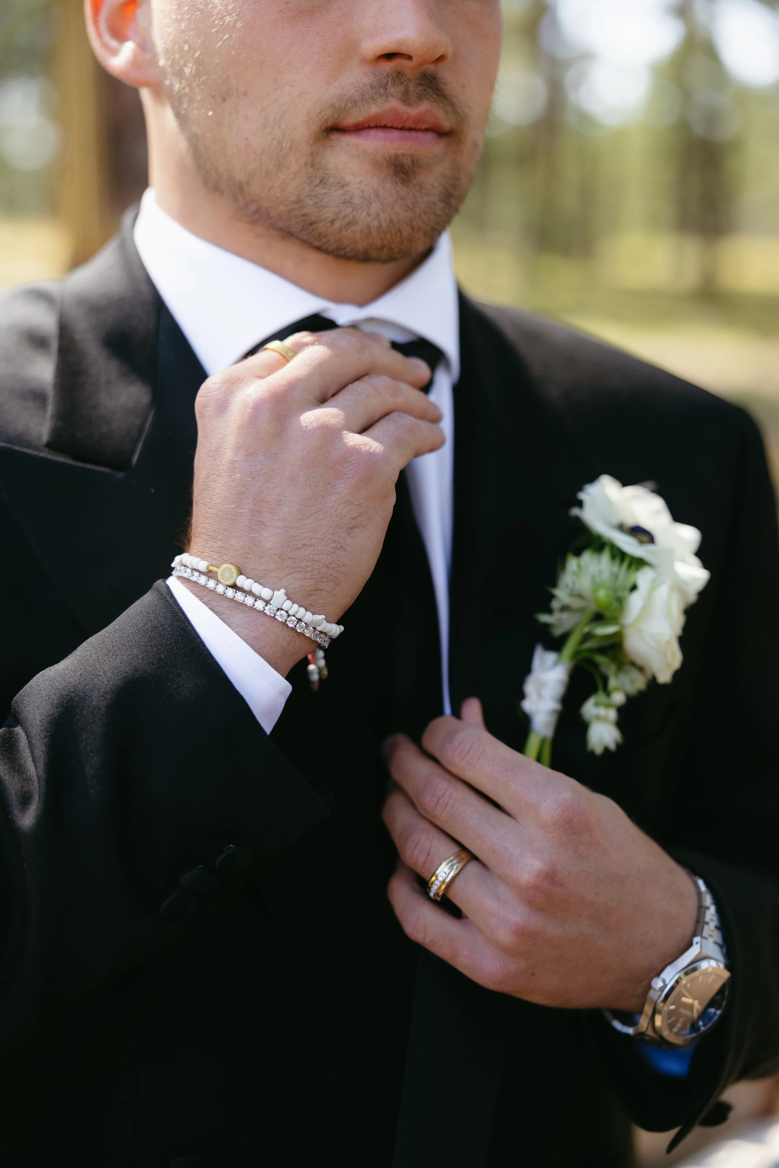 Close-up of a groom in a black tuxedo adjusting his bow tie outdoors, with a white boutonniere on his lapel. The groom is wearing jewelry including a watch and rings.