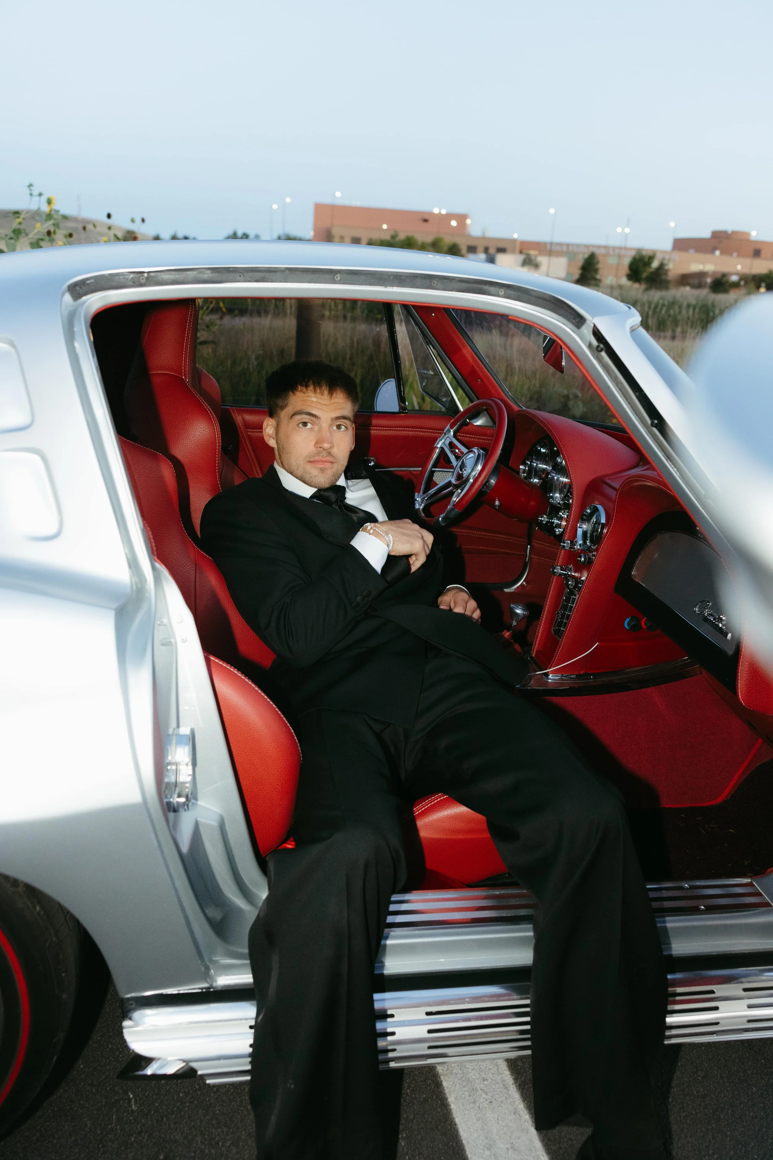Man in formal black tuxedo sitting inside a vintage silver car with red interior, looking at the camera.