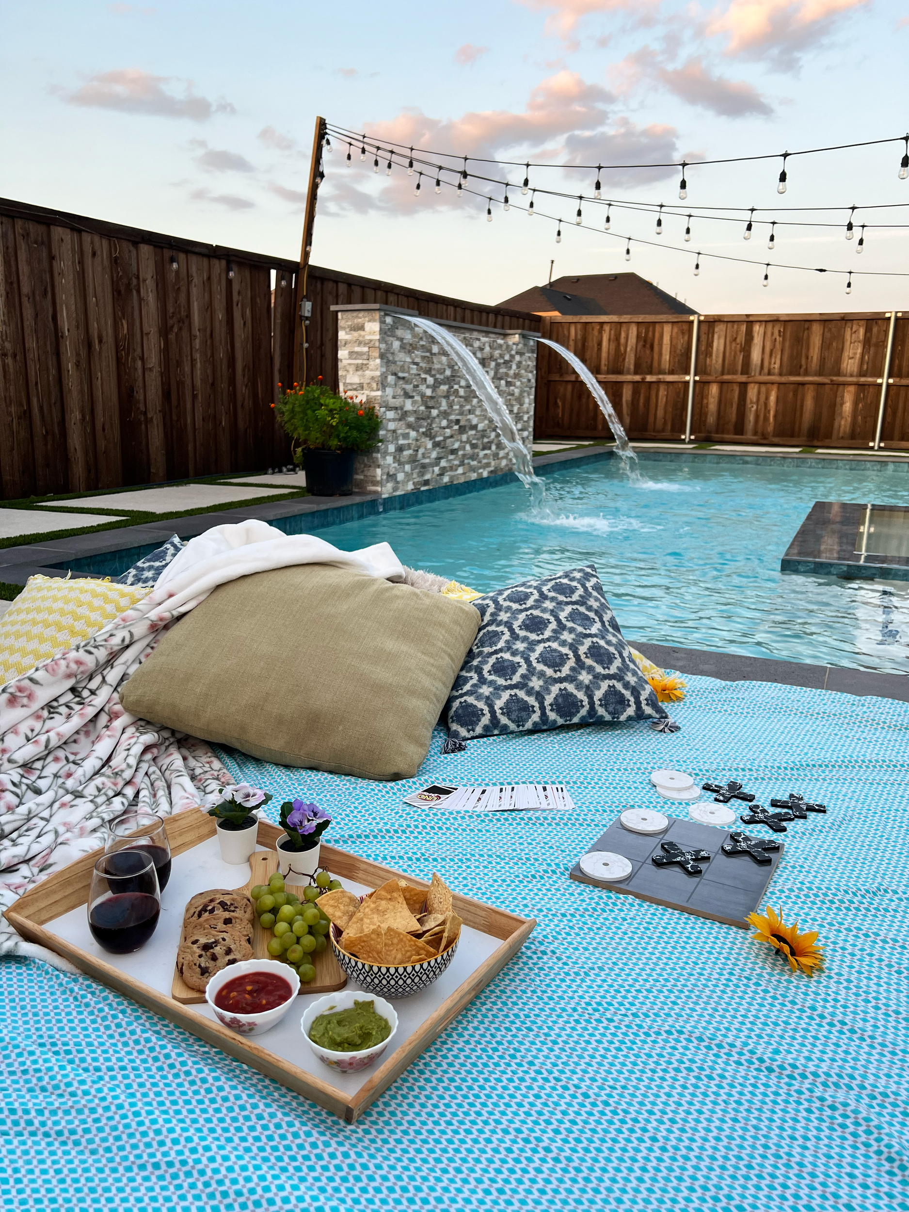Backyard poolside scene with a blanket, pillows, snacks, wine glasses, and play station controllers on a blue and white checkered blanket. In the background, a pool with water features and string lights overhead.