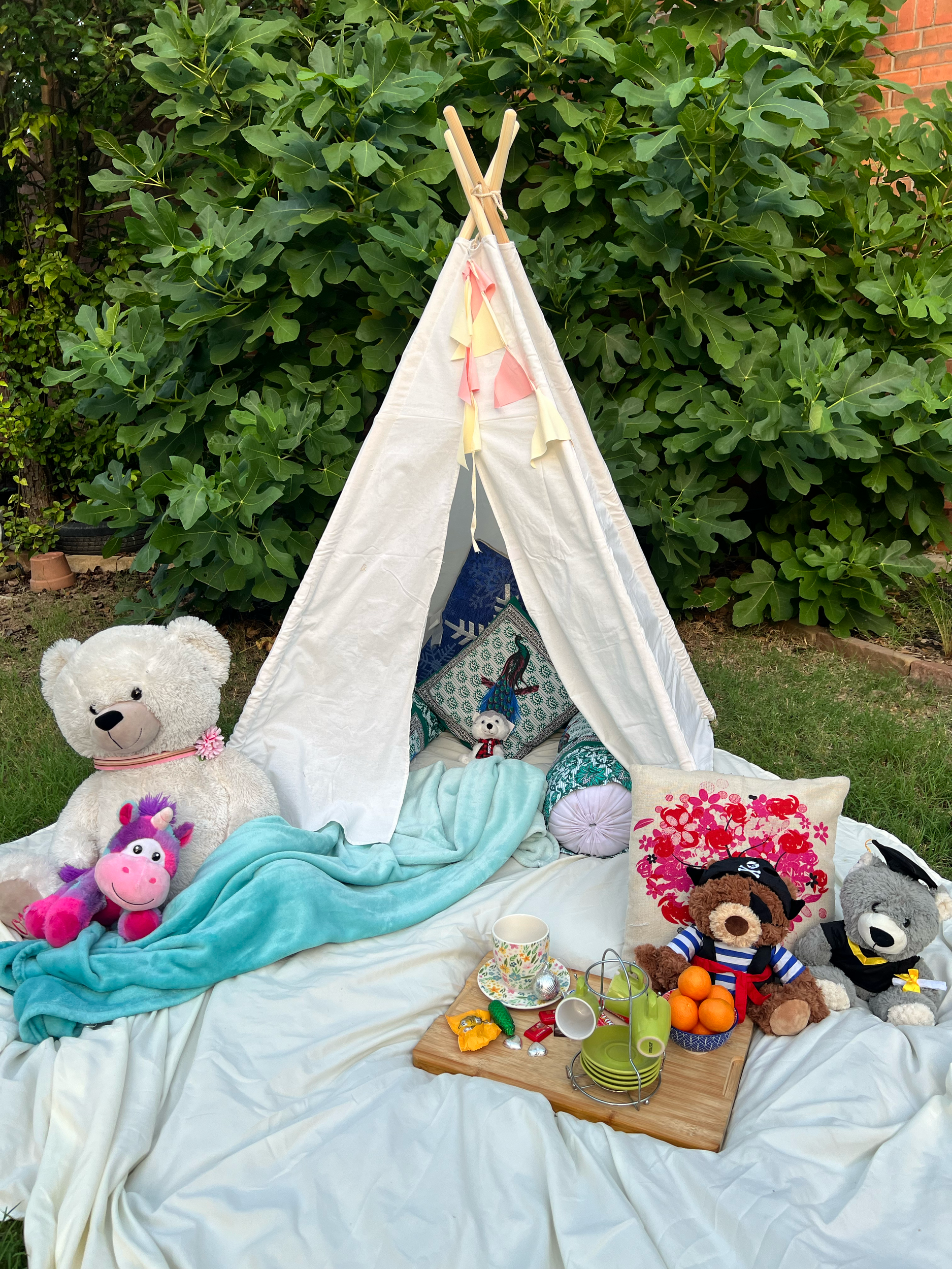 Children's outdoor tea party setup with stuffed animals, a small teepee, and a picnic tray with cups and snacks on the grass.