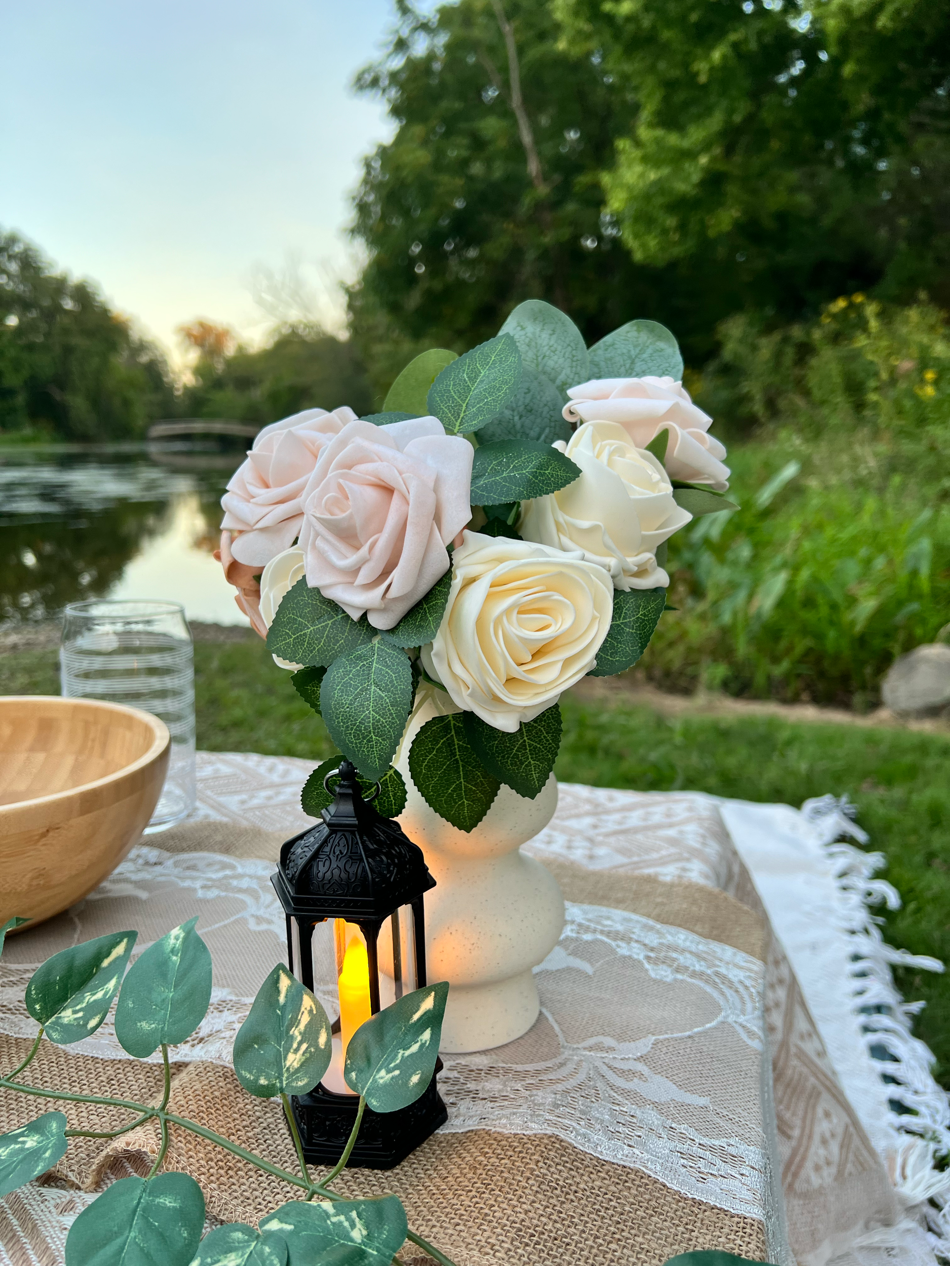 A floral arrangement with white and blush roses, green leaves, a candle, and a small black lantern on a table with lace and burlap tablecloth outdoors with trees and a lake in the background.