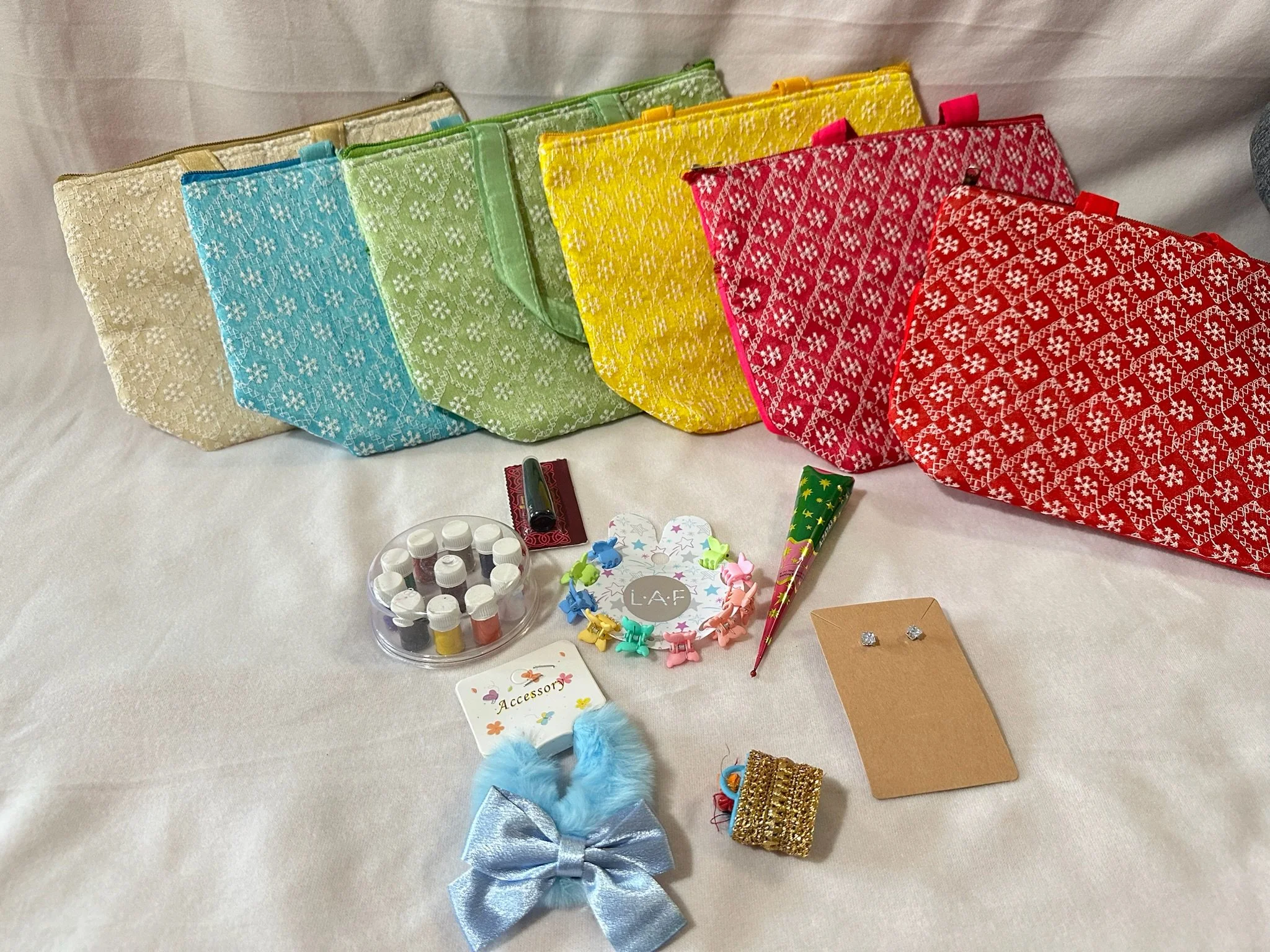 Colorful drawstring bags in beige, blue, green, yellow, pink, and red with white patterns, arranged in a row on a white surface. In front of the bags are small items including a set of nail polish bottles, a round jar of tiny nail polish bottles, colorful star-shaped clips, a small gold box, a brown card with earrings, a light blue fluffy bow with a silver ribbon, a small decorated cone, and a card labeled "Accessory."
