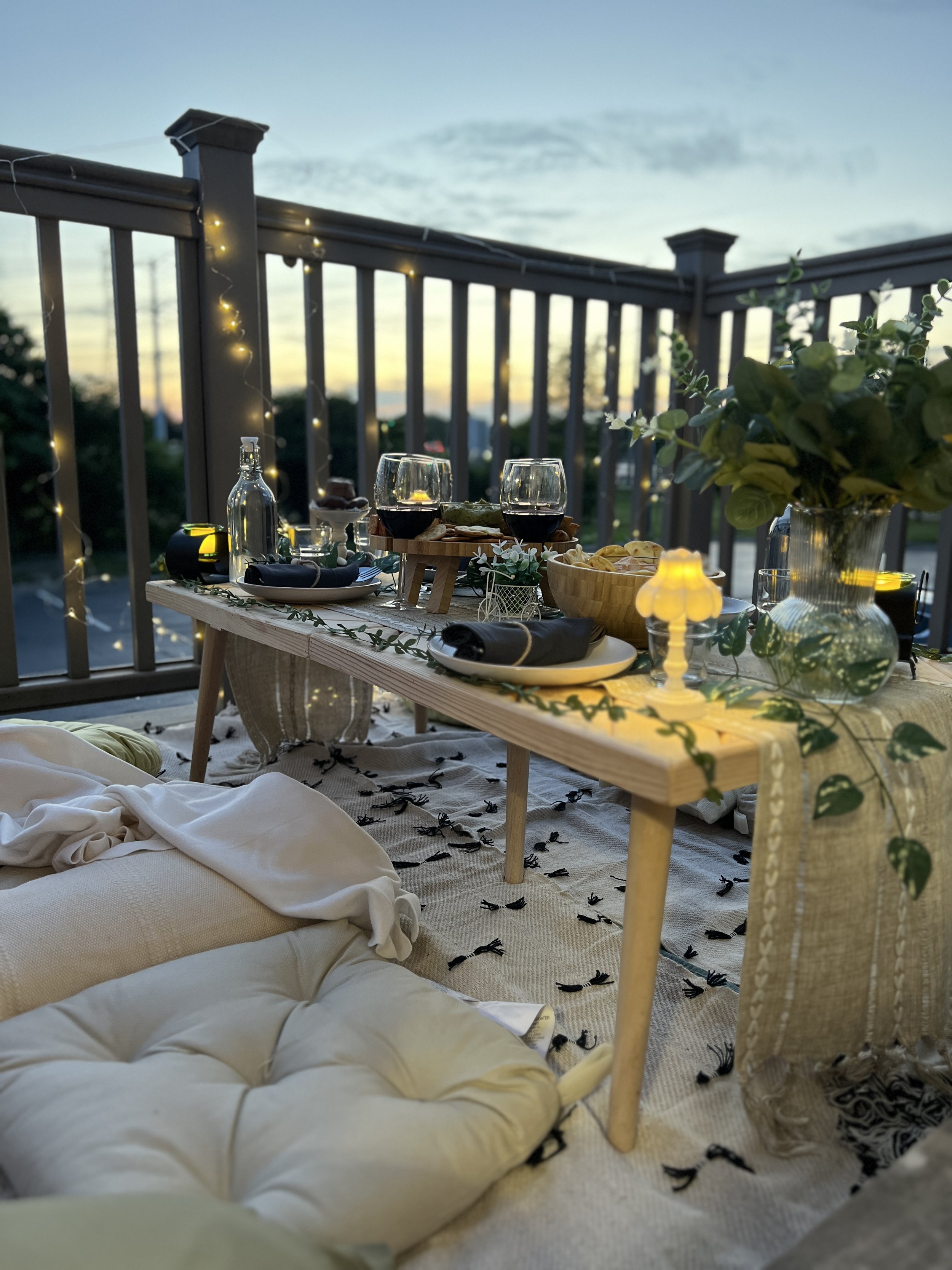 A cozy outdoor dinner setup on a balcony at sunset, featuring a wooden table with wine glasses, a bowl of chips, and a plant in a glass vase. String lights and a blanket-covered cushion are also visible.