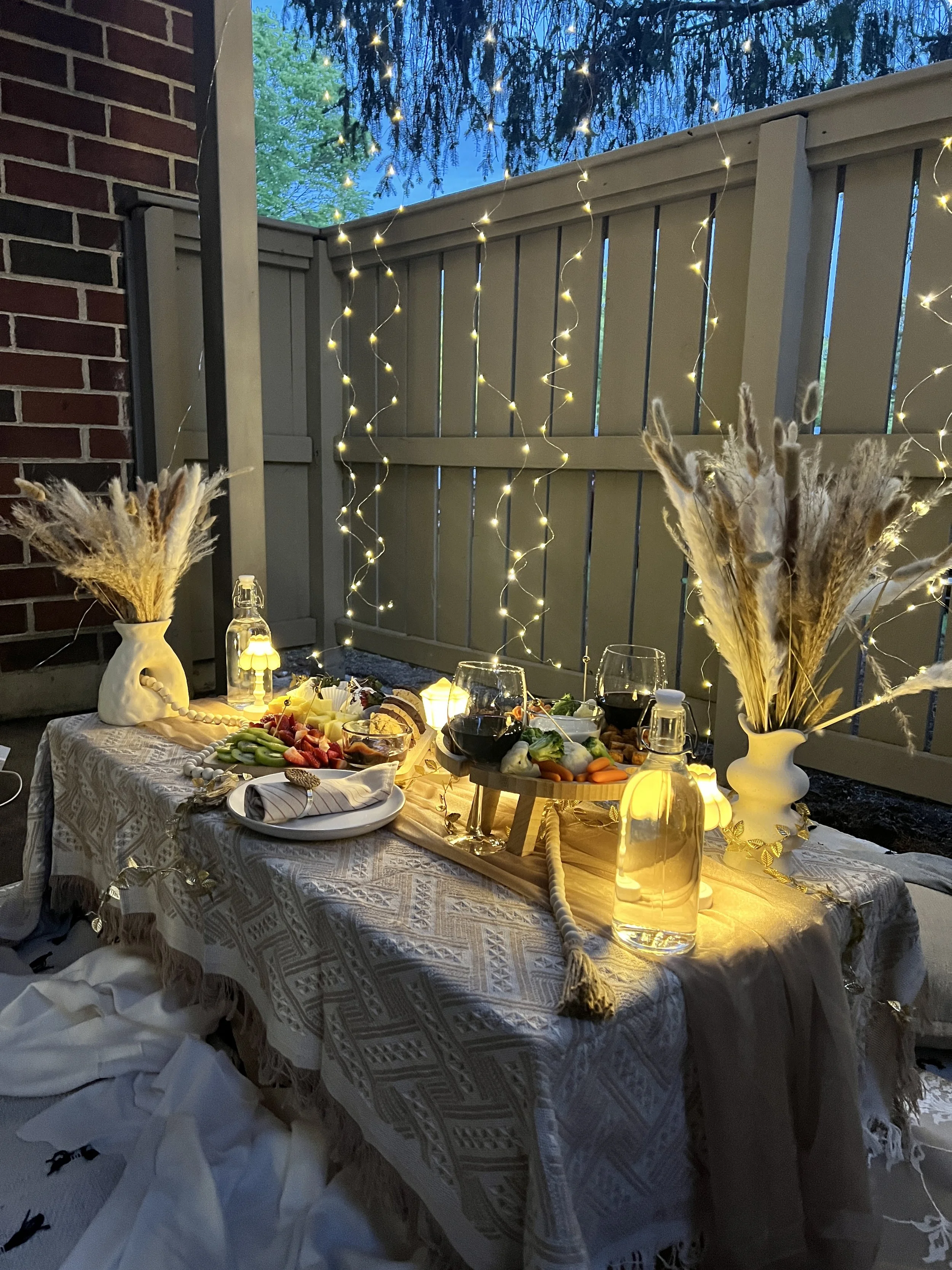 Outdoor dining table decorated with pampas grass in vases, candles, wine glasses, and a fruit platter, illuminated with string lights against a fence at dusk.
