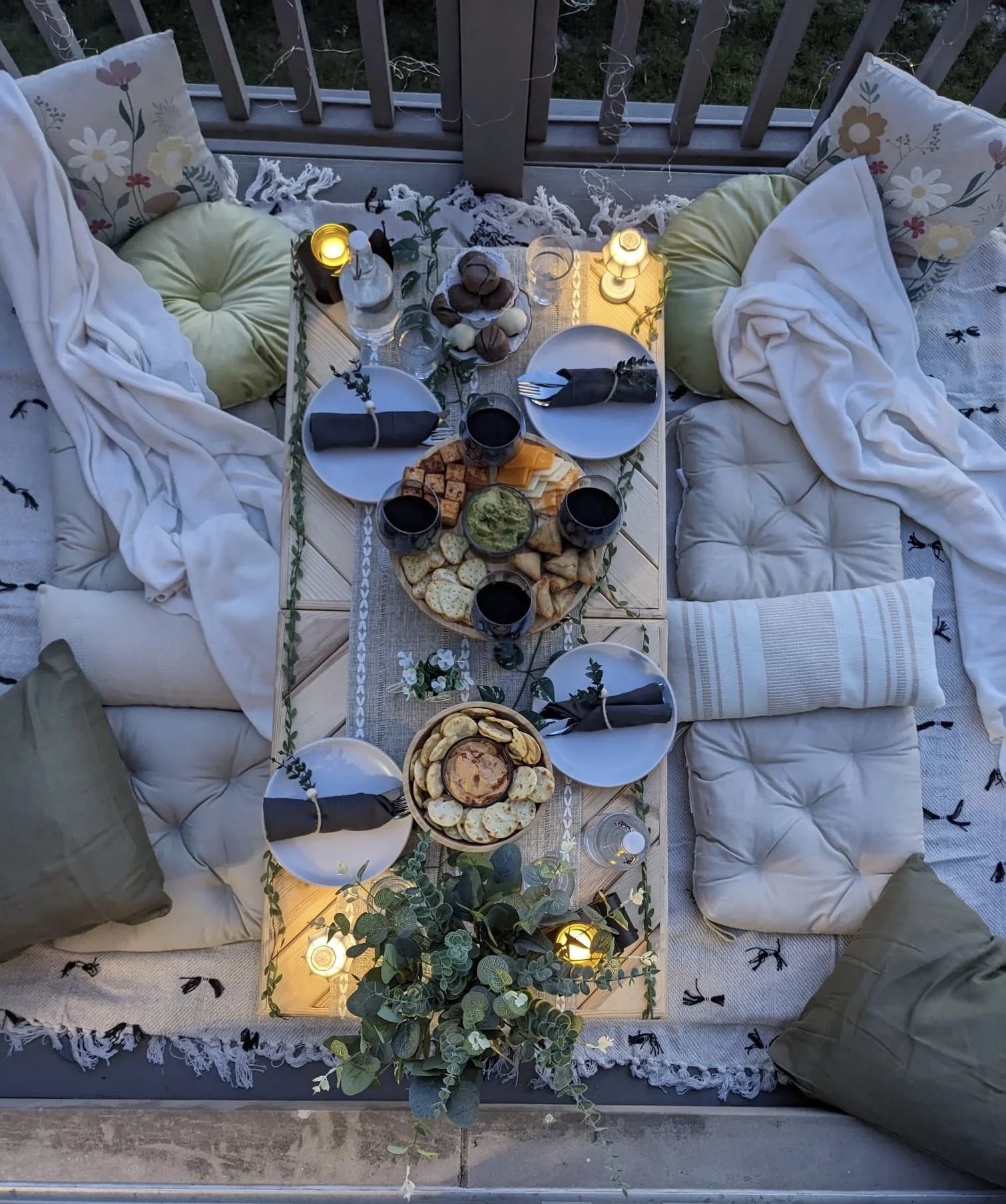 Overhead view of a cozy outdoor dining setup on a low table with plates, glasses of red wine, a variety of snacks including cheese, crackers, bread, and dips, surrounded by cushions and pillows on the floor, decorated with small candles and greenery.