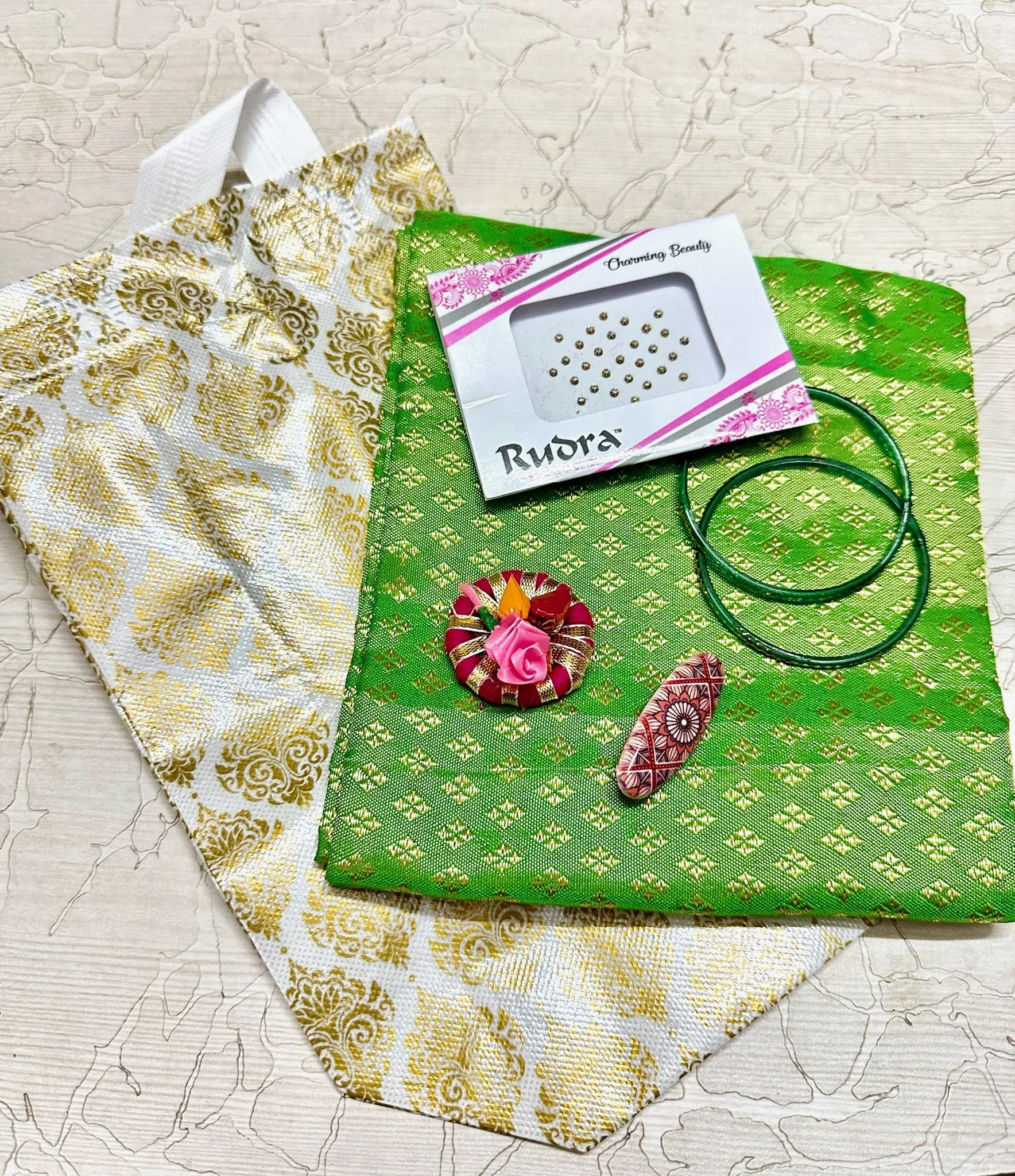An assortment of jewelry and fabric items, including green and white bangles, a decorative pink and gold ring, a pink and white patterned finger ring, a box of small gold and silver beads, all placed on green and white patterned fabric with a gold and white patterned cloth beneath.