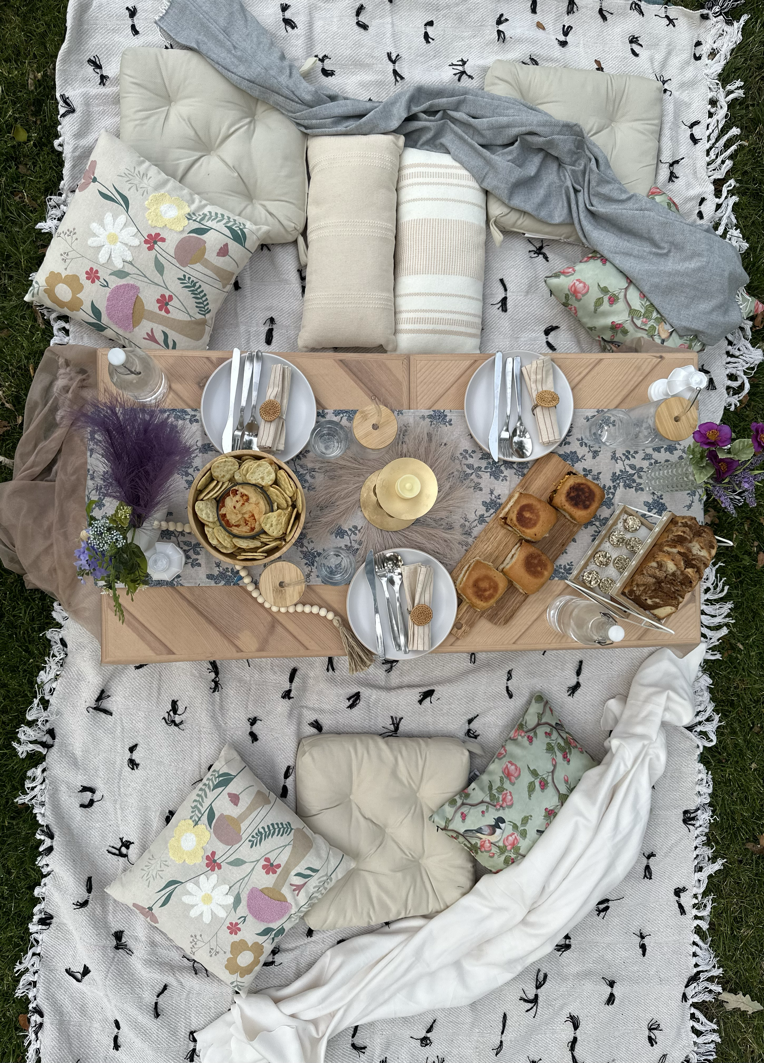 A picnic setup on a blanket with pillows, cushions, and a table with food and drinks on a grassy area.
