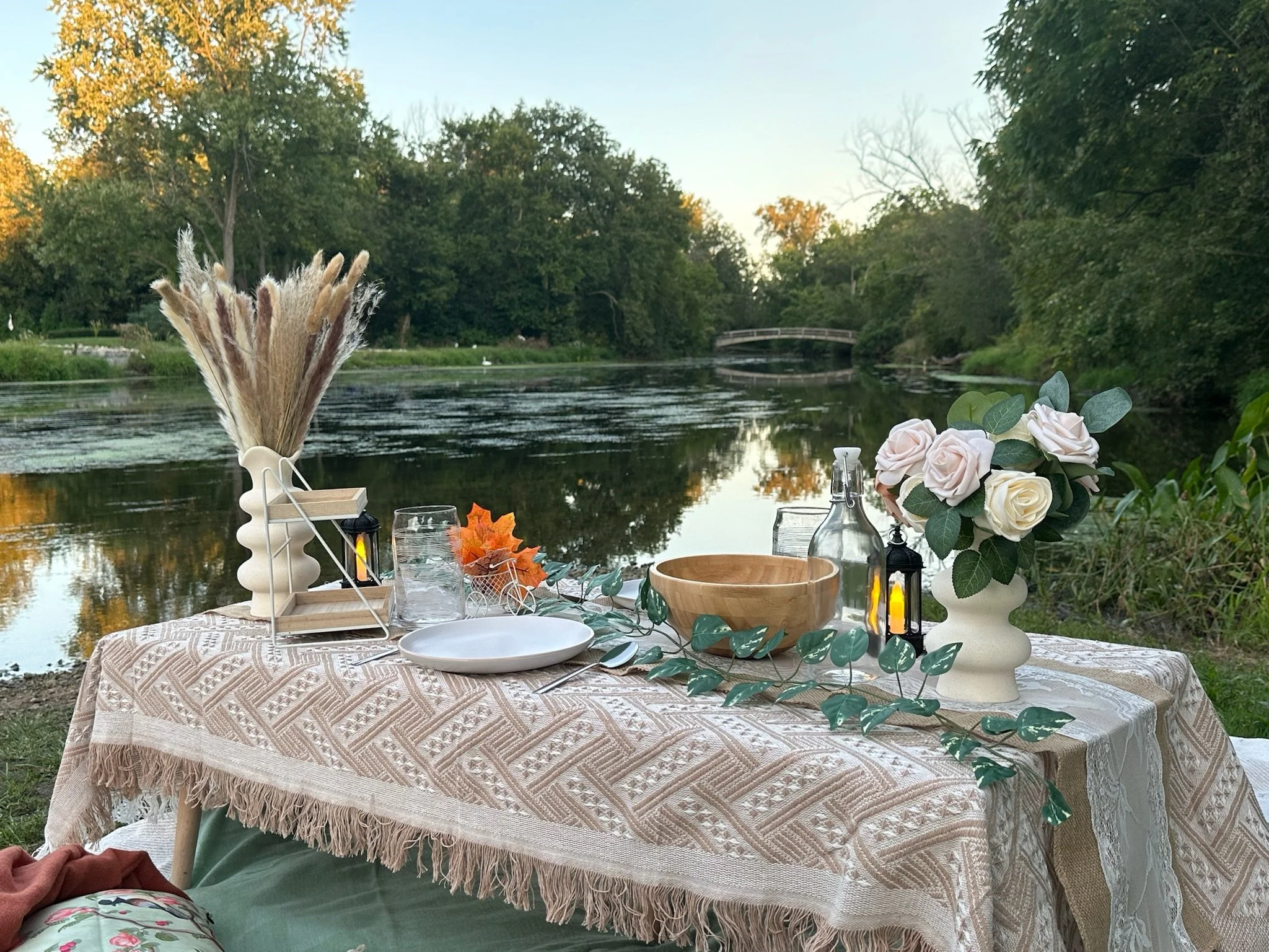 A lakeside outdoor table set for a meal with decorative vases, candles, plates, and a wooden bowl, against a backdrop of a calm river, trees, and a small bridge at sunset.