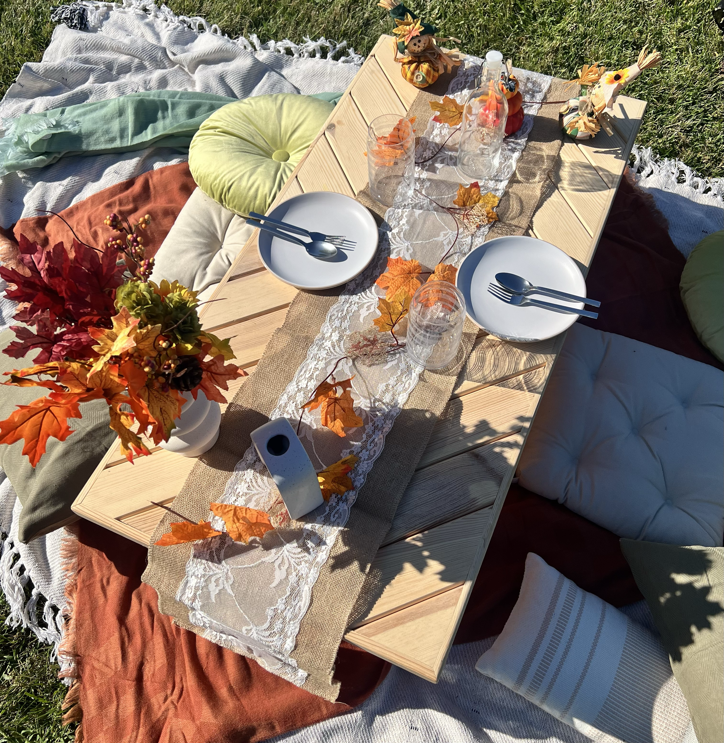Outdoor autumn-themed dining setup with a wooden table decorated with fall leaves, a vase of colorful fall flowers, white plates with utensils, glasses, and small decorative scarecrows, surrounded by cushions and blankets on grass.