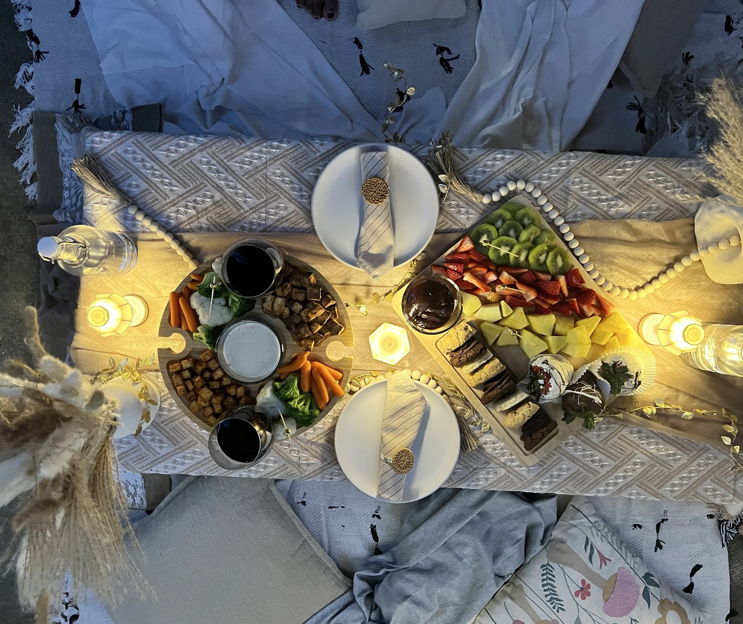 Table set for a meal with plates, cutlery, wine glasses, candles, and a variety of food including fruits, vegetables, cheeses, and desserts, decorated with string lights and candles in a cozy setting.