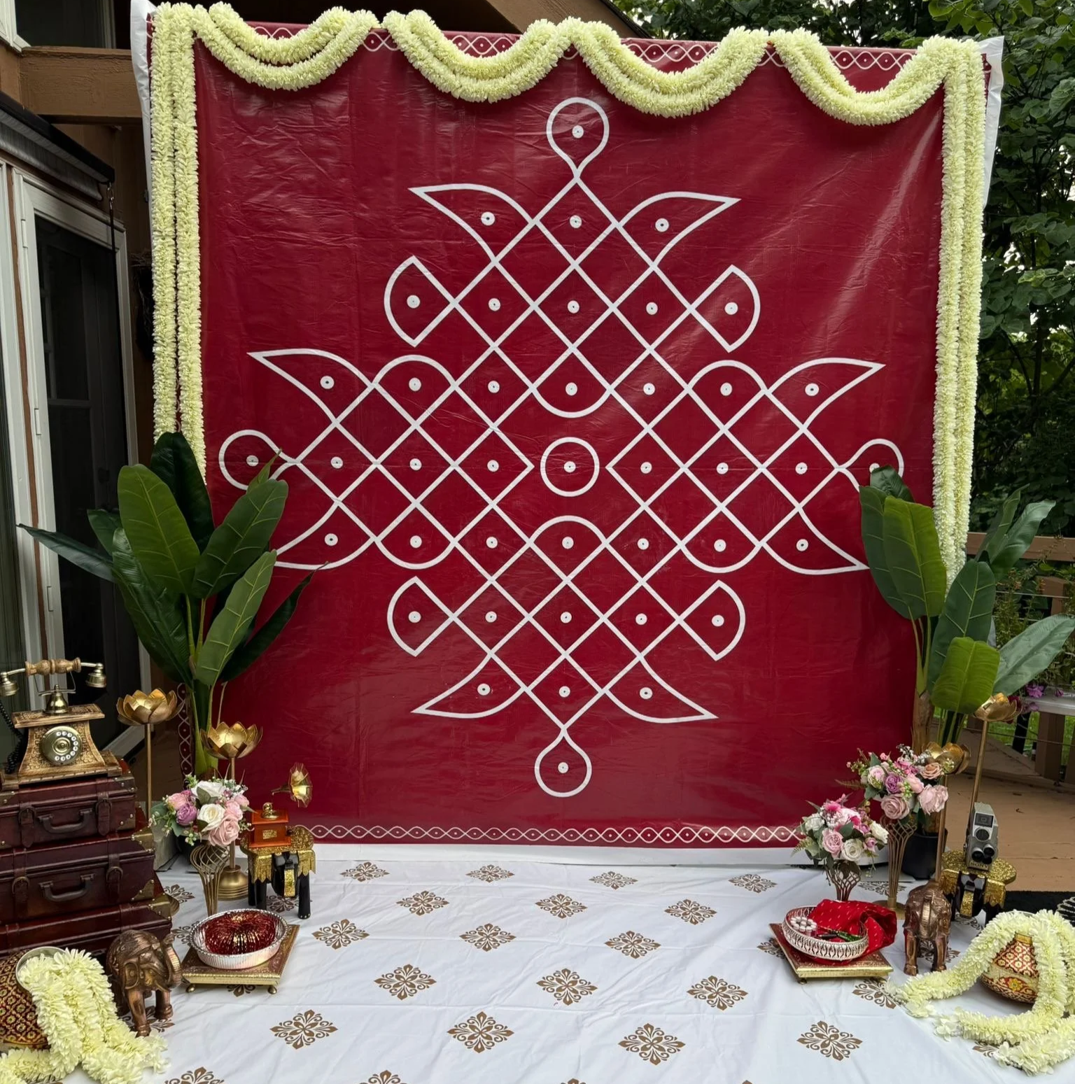 Decorative Indian wedding mandap with a red backdrop, white traditional geometric design, floral garlands, green leaves, and various traditional items including elephant figurines and flower arrangements.
