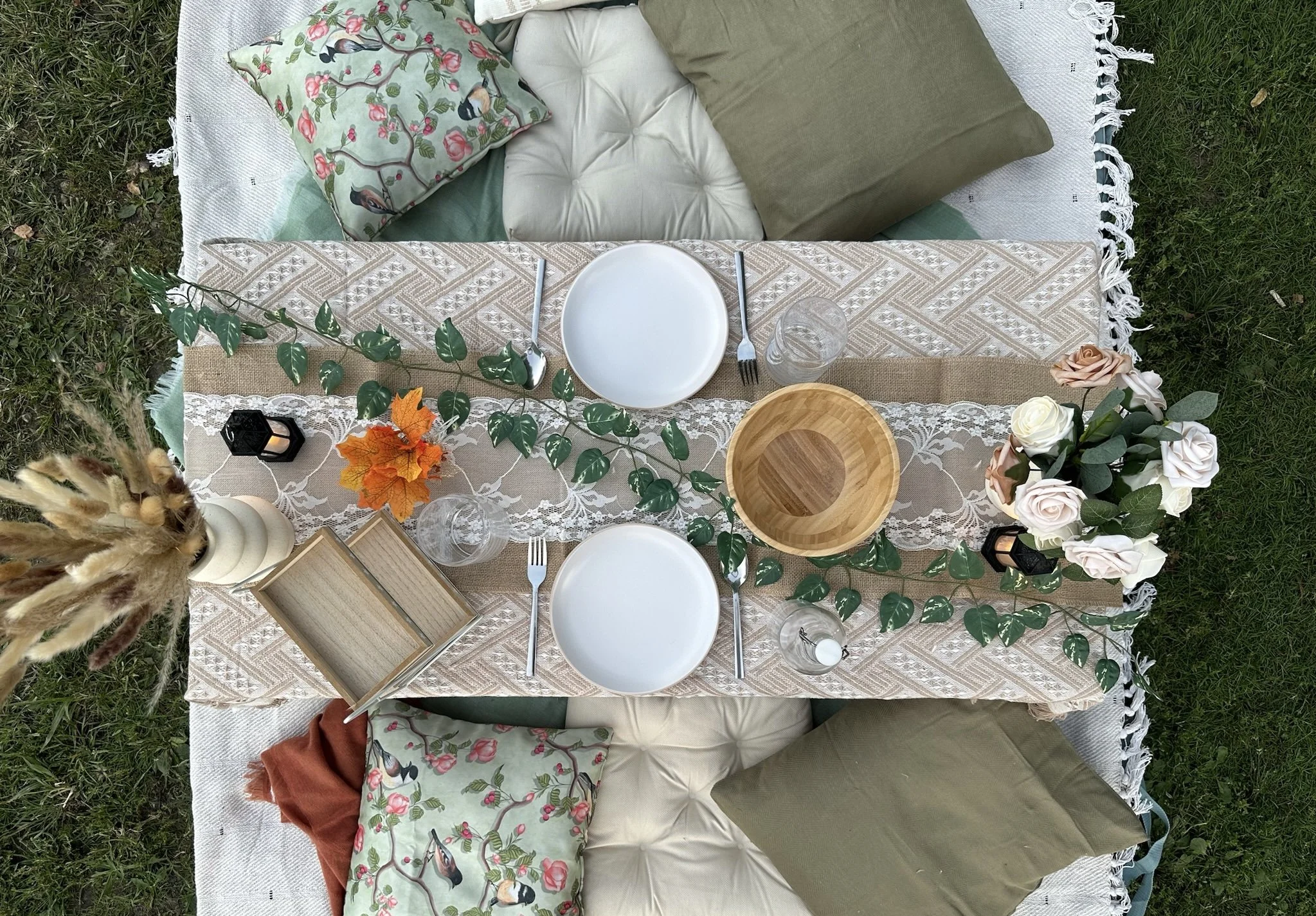 Outdoor picnic table set with plates, glasses, cutlery, and a centerpiece of flowers and greenery on a patterned tablecloth, surrounded by cushions and pillows on the grass.