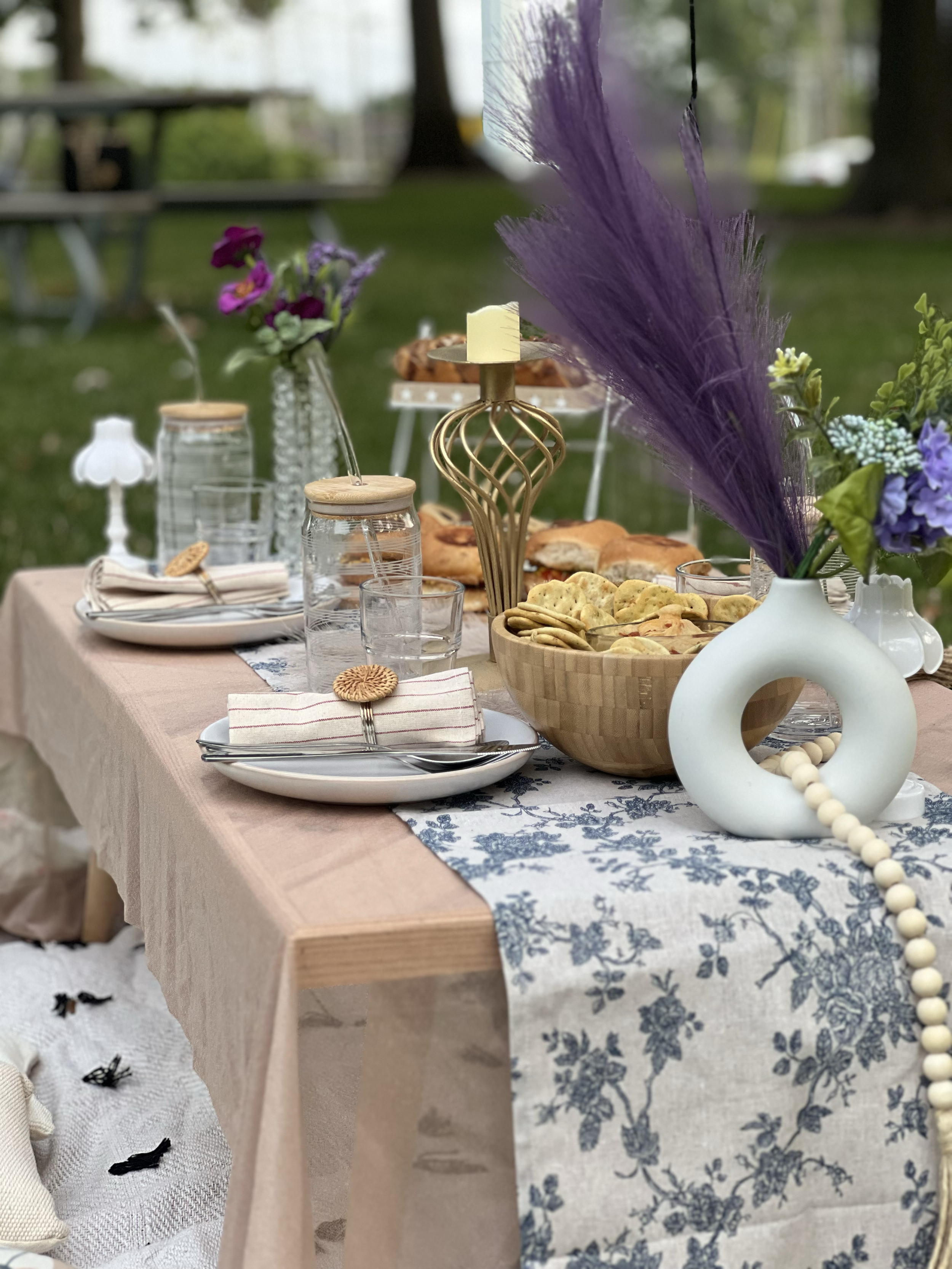 Decorative outdoor table set with flowers, snacks, and tableware on a beige tablecloth and floral runner, with trees and park in the background.