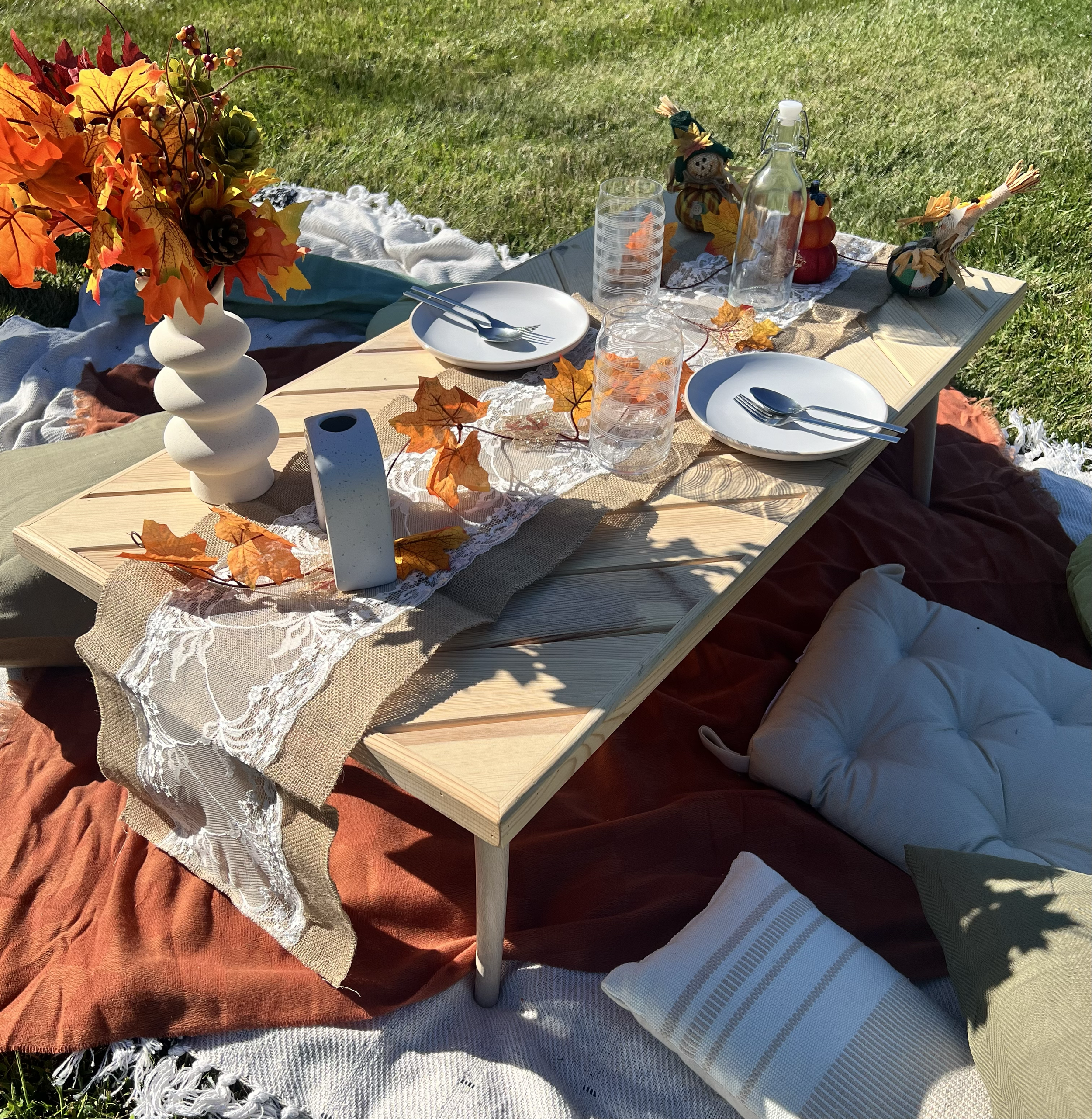 A picnic table set on the grass with fall-themed decorations, plates, glasses, and pillows for seating.