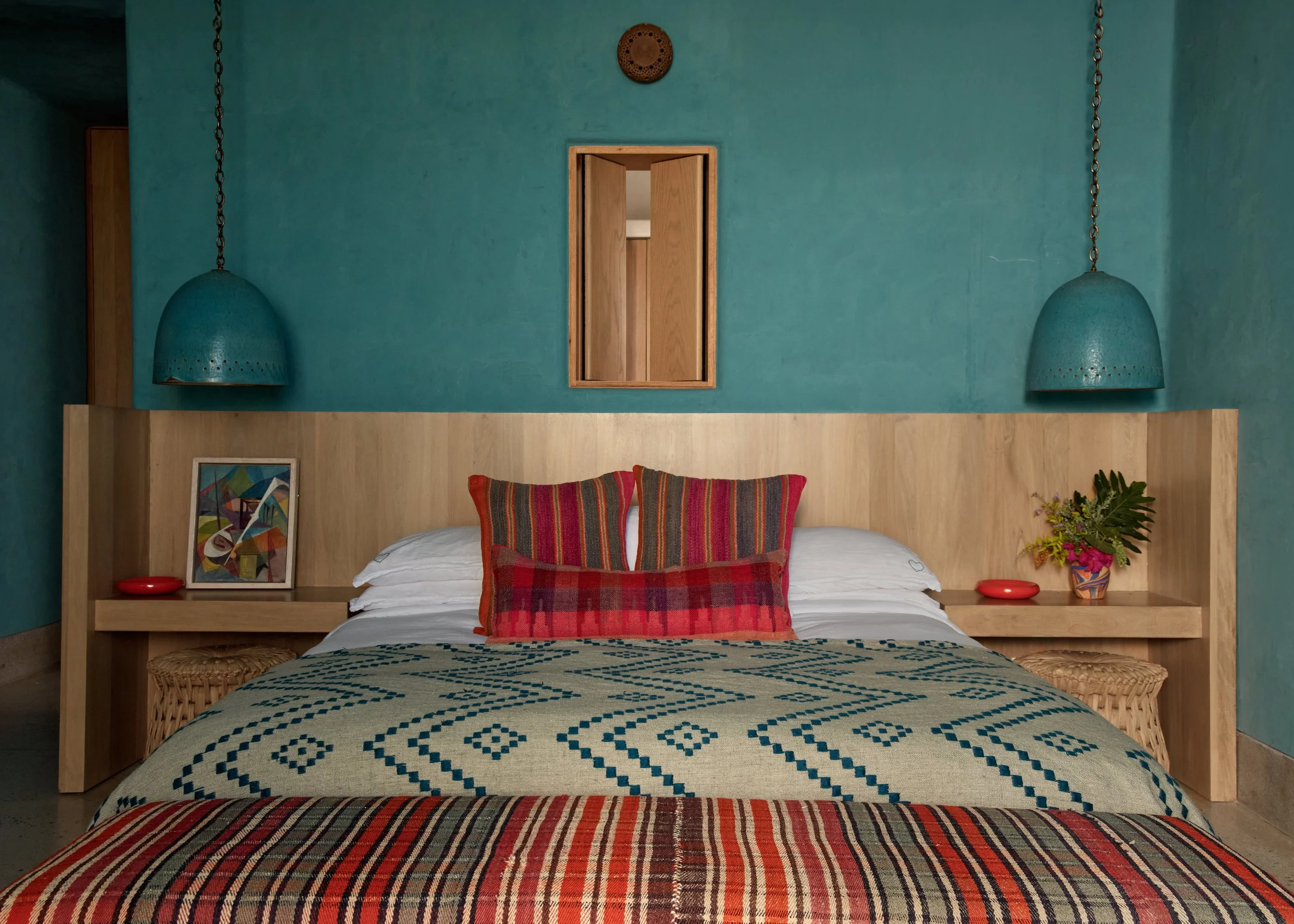 A bedroom with a wooden headboard, two red and black plaid pillows, a colorful abstract painting on the wall, a nightstand with a red dish, a colorful vase with flowers, and teal lamps hanging from the ceiling. The wall behind the bed is painted teal, and the lower part has wood paneling.