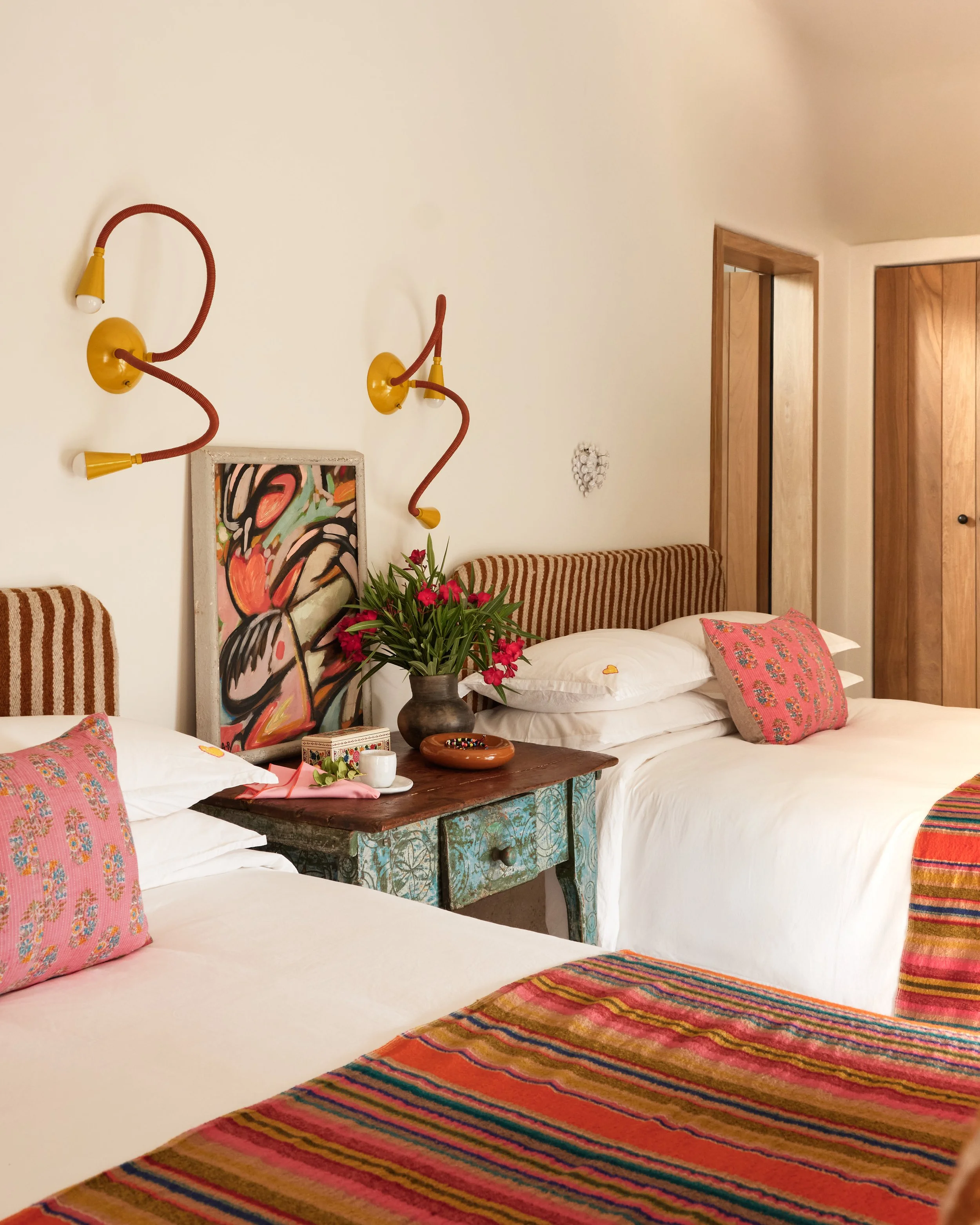 A bedroom with two beds with white linens and pink flower-patterned pillows. There is a rustic wooden nightstand with a potted plant, a bowl, and small decorative items. The wall behind the beds has yellow wall-mounted lights shaped like flexible goo
