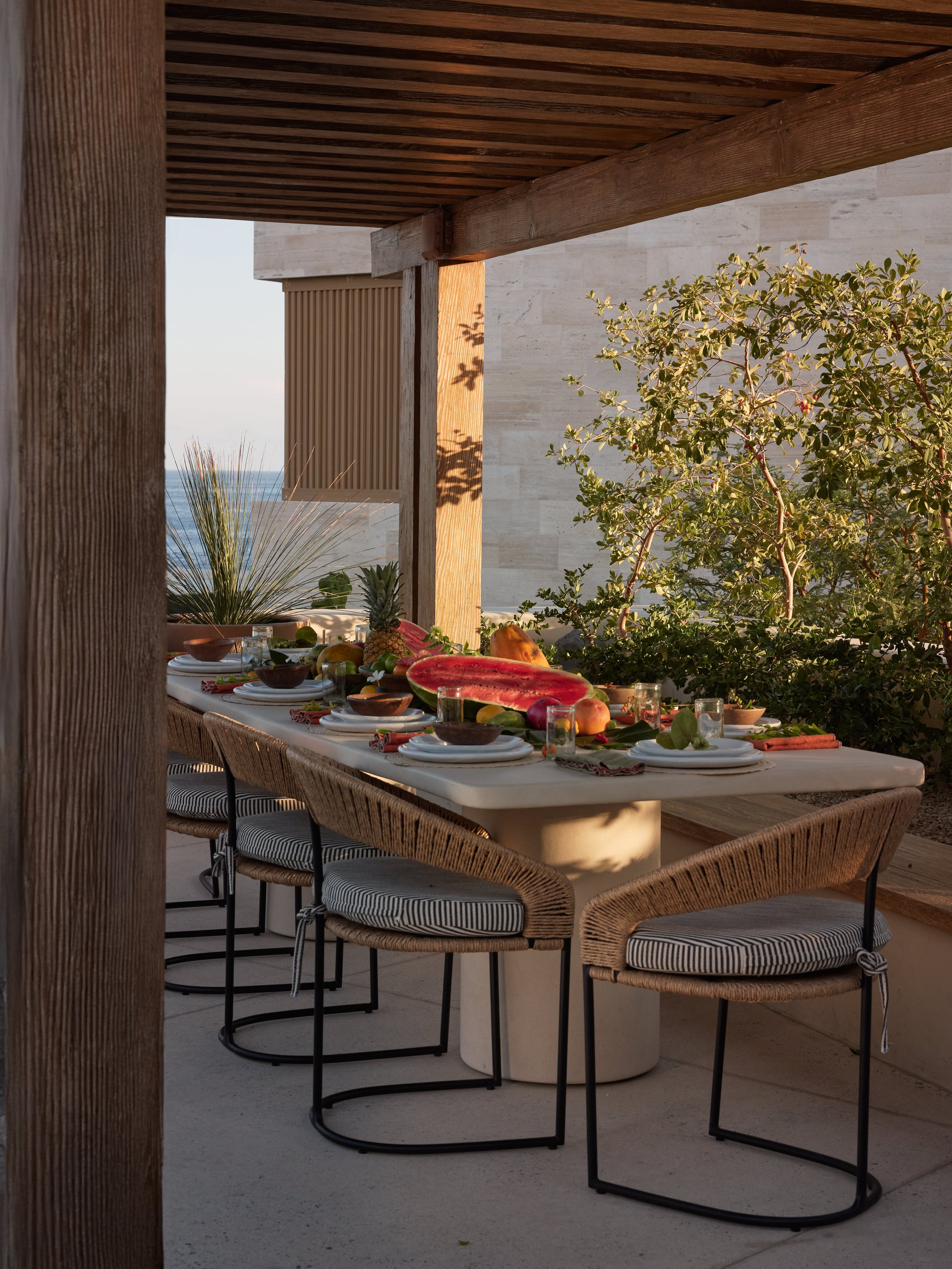 Outdoor dining table set with plates, bowls, and large slices of watermelon, surrounded by chairs with striped cushions, under a wooden pergola with greenery in the background.