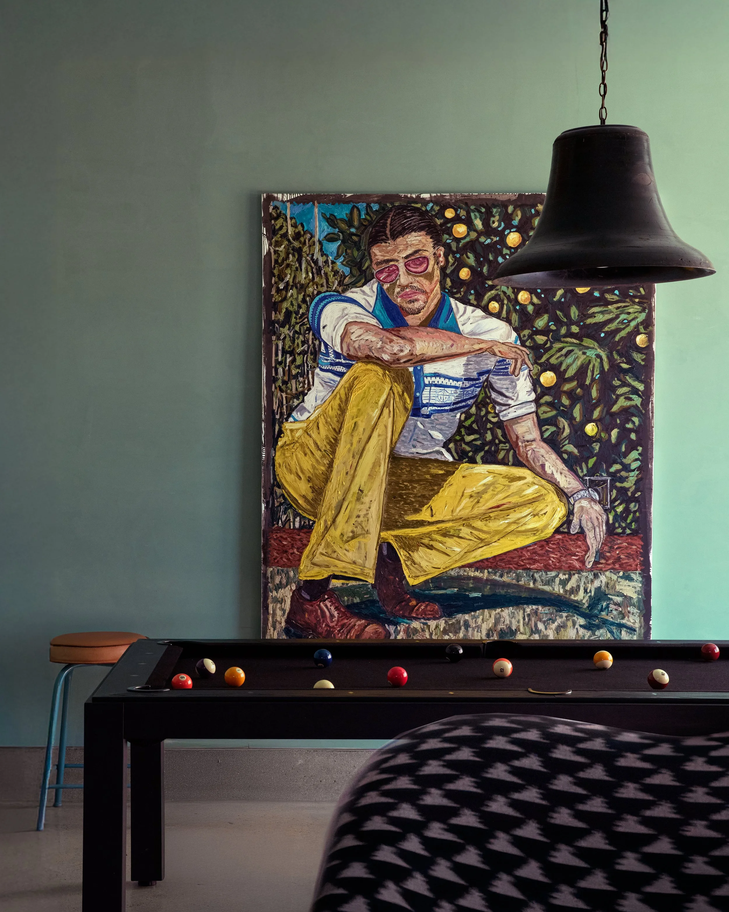 Painting of a man with dark hair and sunglasses, seated with legs crossed, wearing a white shirt with blue details and yellow pants, set against a background of green foliage and yellow fruit. The photo also shows a black pool table with billiard bal