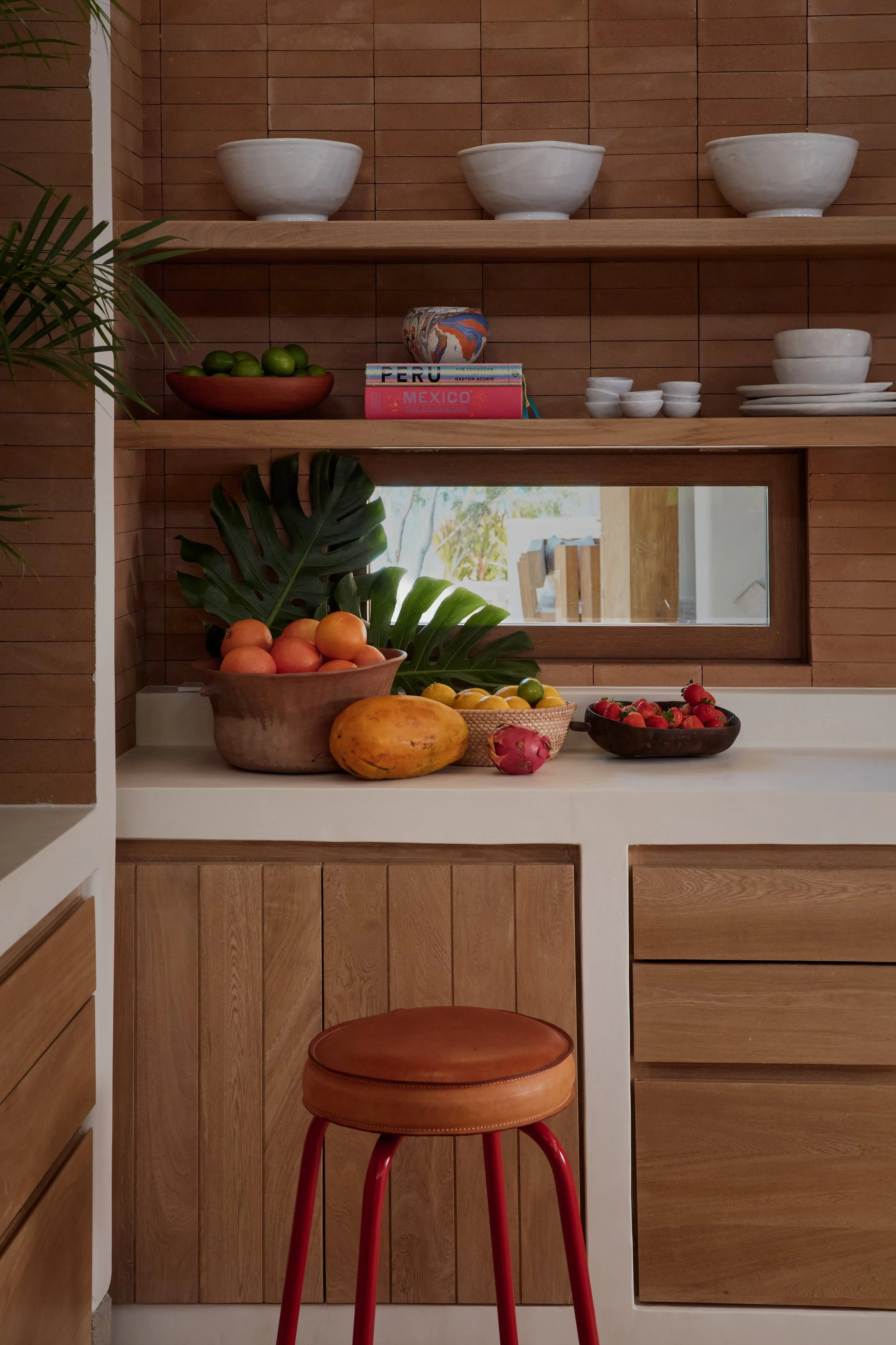 A kitchen shelf with white bowls and smaller dishes, a decorative mug, and books about Peru and Mexico. Below the shelf, a counter with a bowl of oranges, a papaya, and strawberries, with a window reflecting the outdoor scenery. A red stool with a round leather top is in front of the counter.