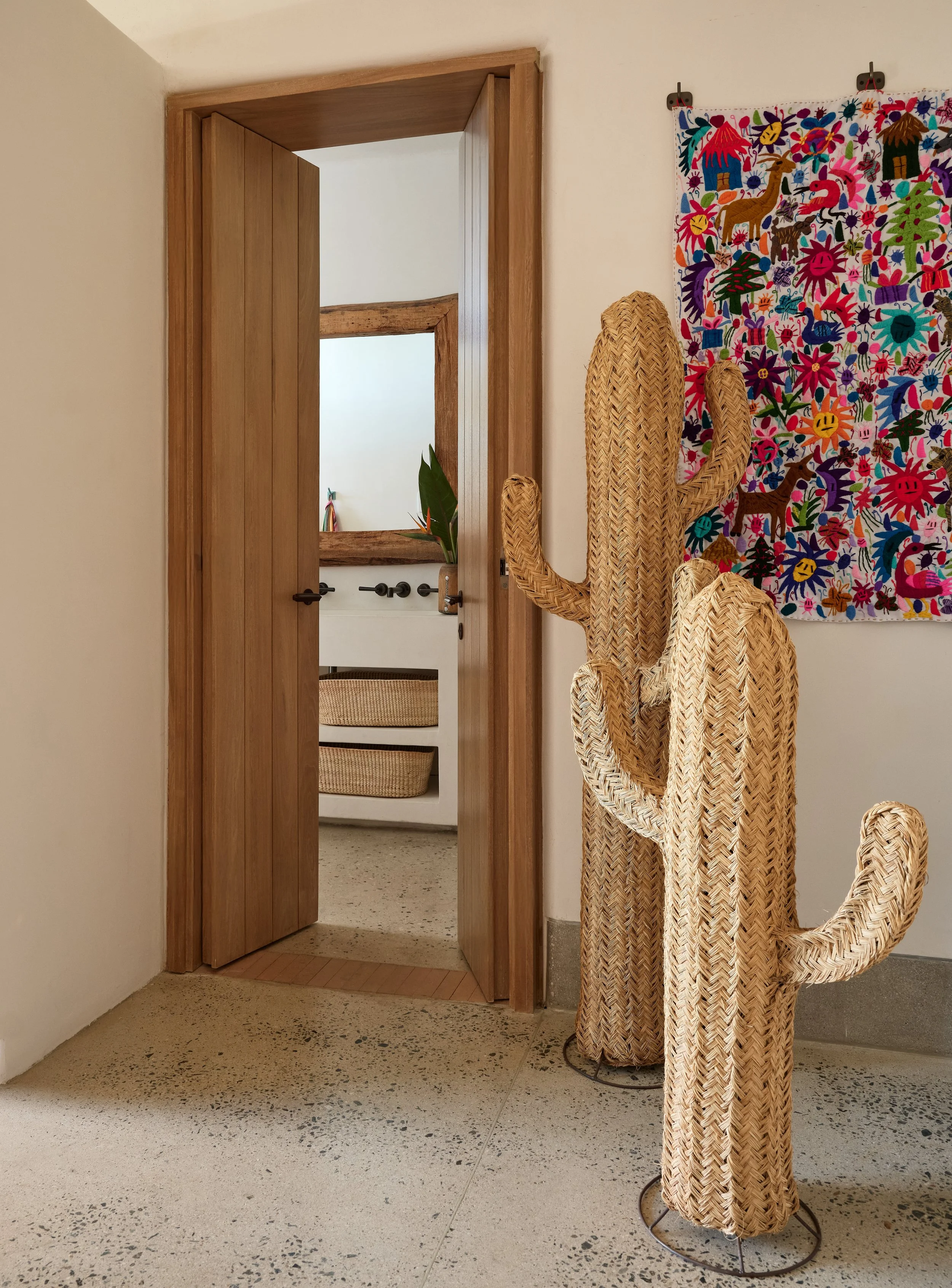 Woven cactus sculpture in front of colorful embroidered wall hanging, seen through an open wooden door in an indoor setting.