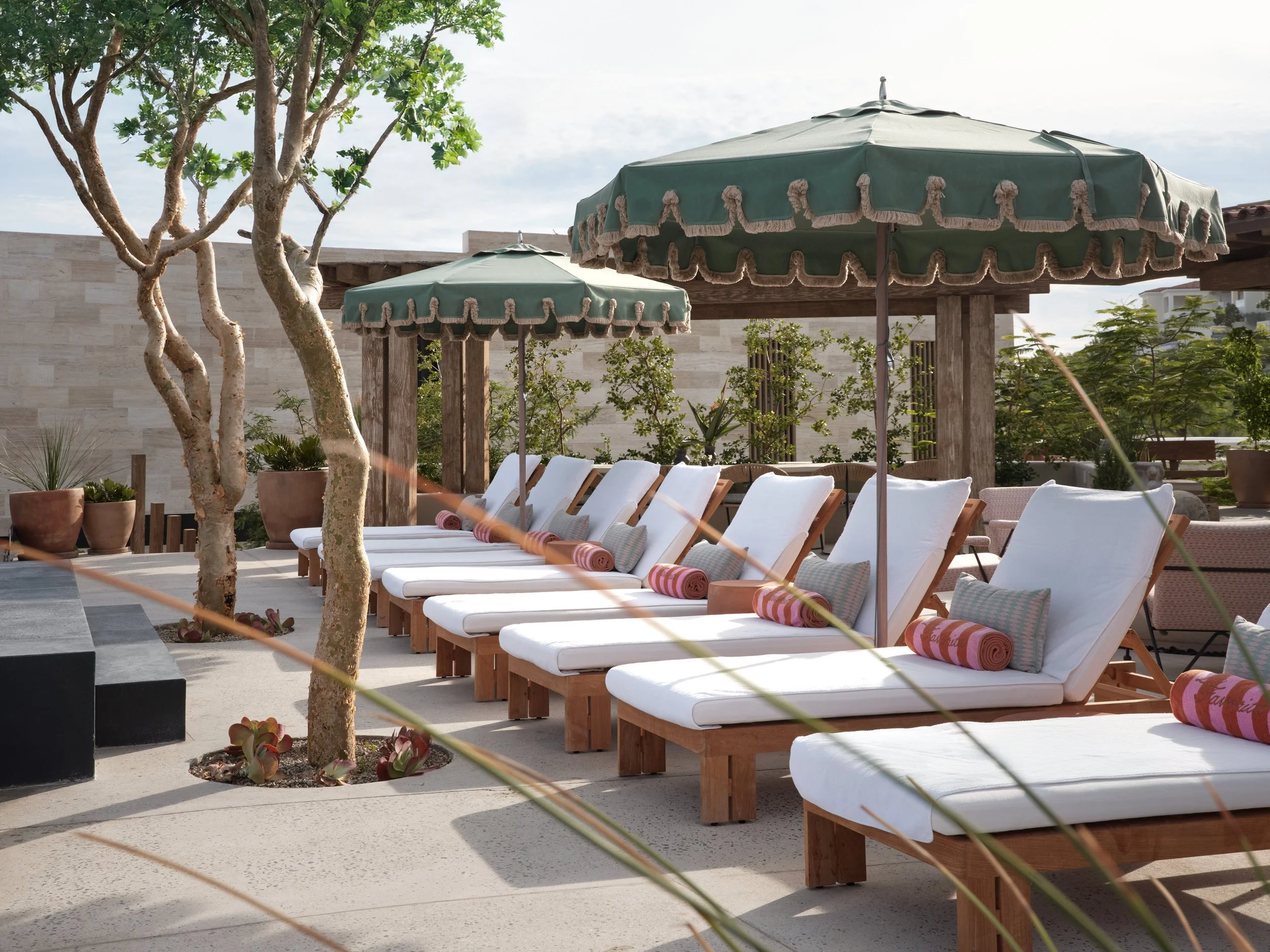 Outdoor patio with white lounge chairs, green umbrellas with beige fringe, potted plants, trees, and a stone wall under a partly cloudy sky.