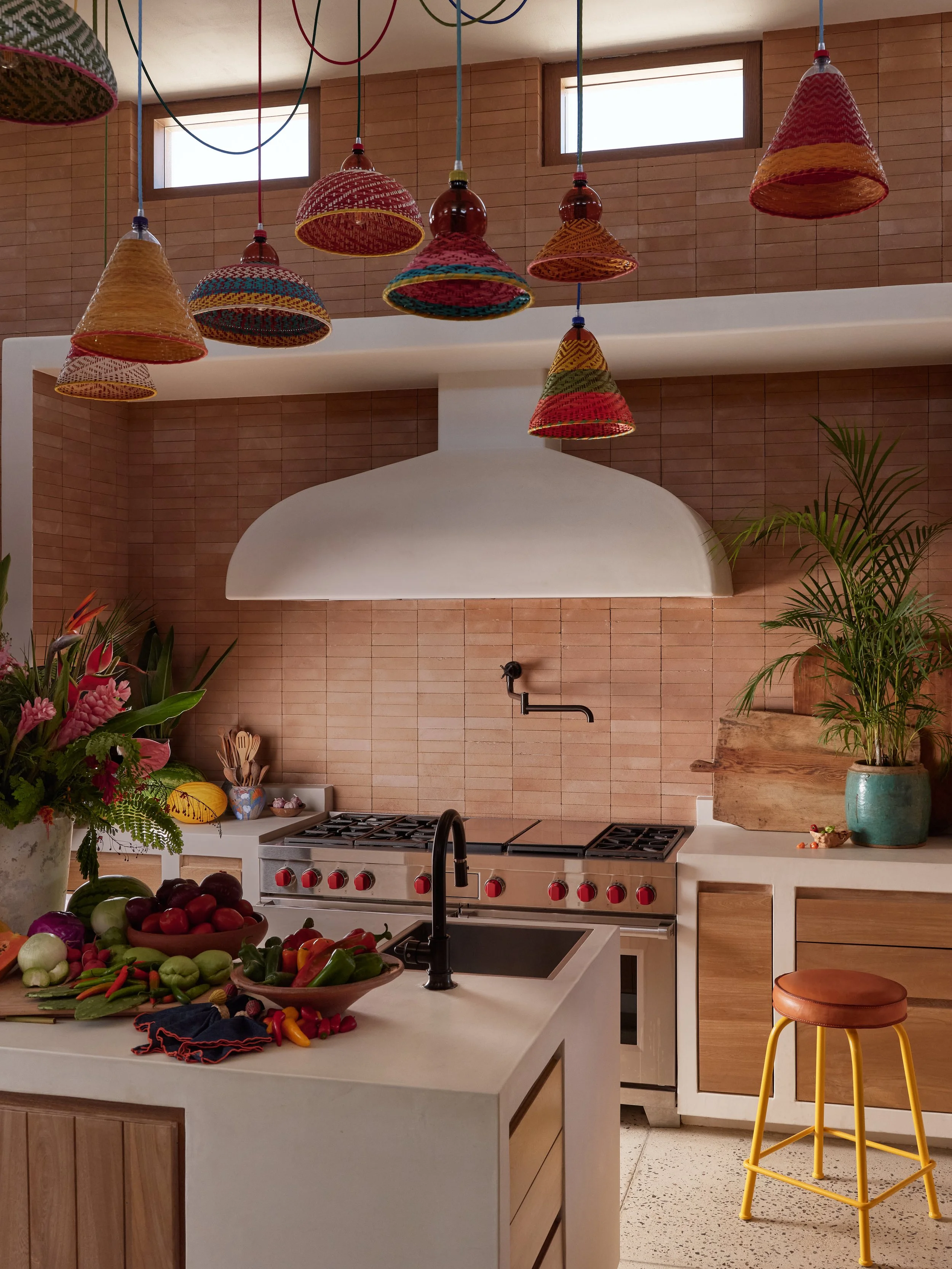 A modern kitchen with a white island counter showcasing a large assortment of fresh vegetables, including radishes, peppers, and leafy greens. In the background, a stove with red knobs is visible beneath a brick wall, which also features a black faucet and a white range hood. Wooden cabinetry and large windows are present, along with colorful woven pendant lights hanging from the ceiling and a green potted plant on the right.