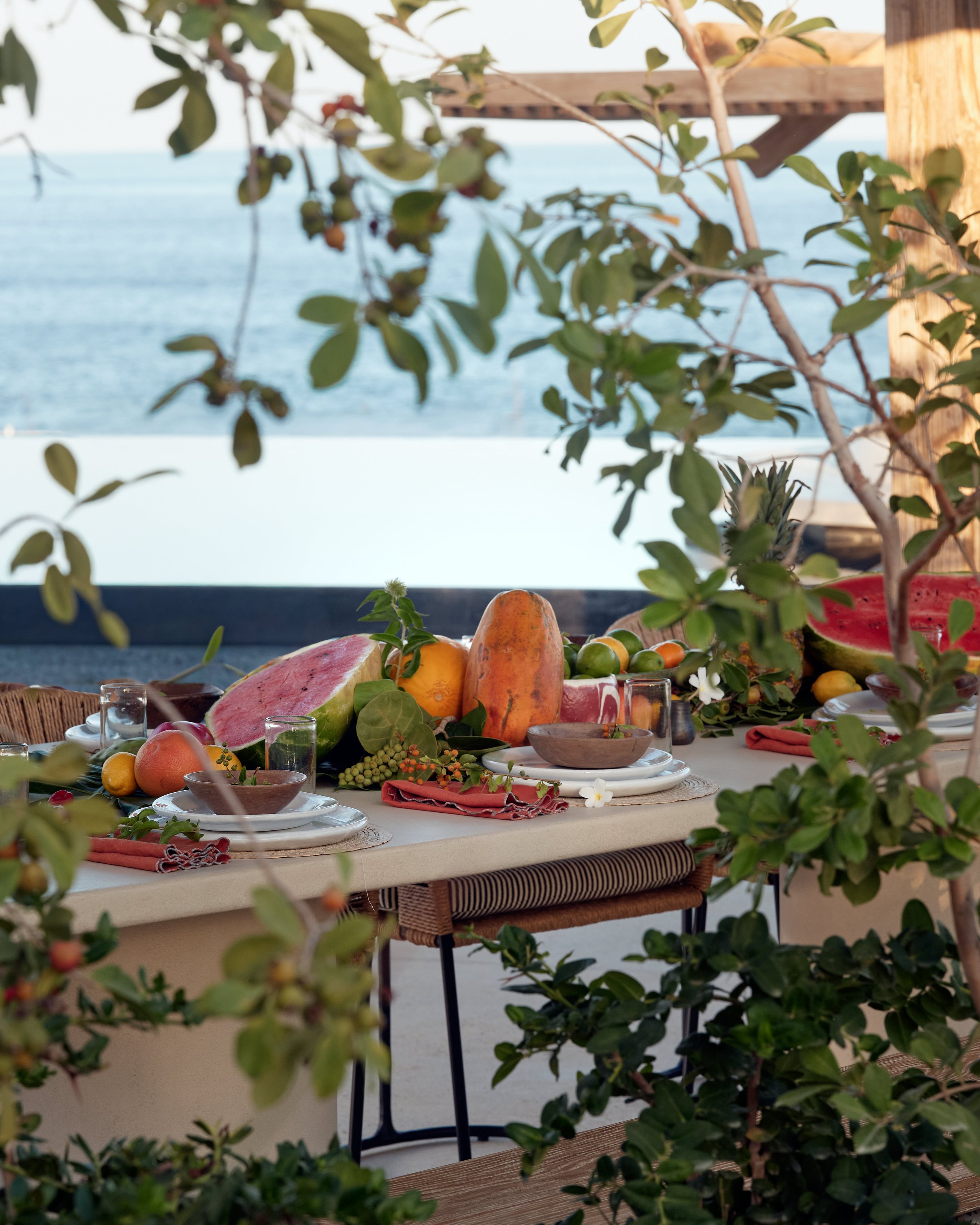 A table set outdoors with watermelons, papayas, lemons, small bowls, glasses, and tableware, decorated with greenery, overlooking the ocean.