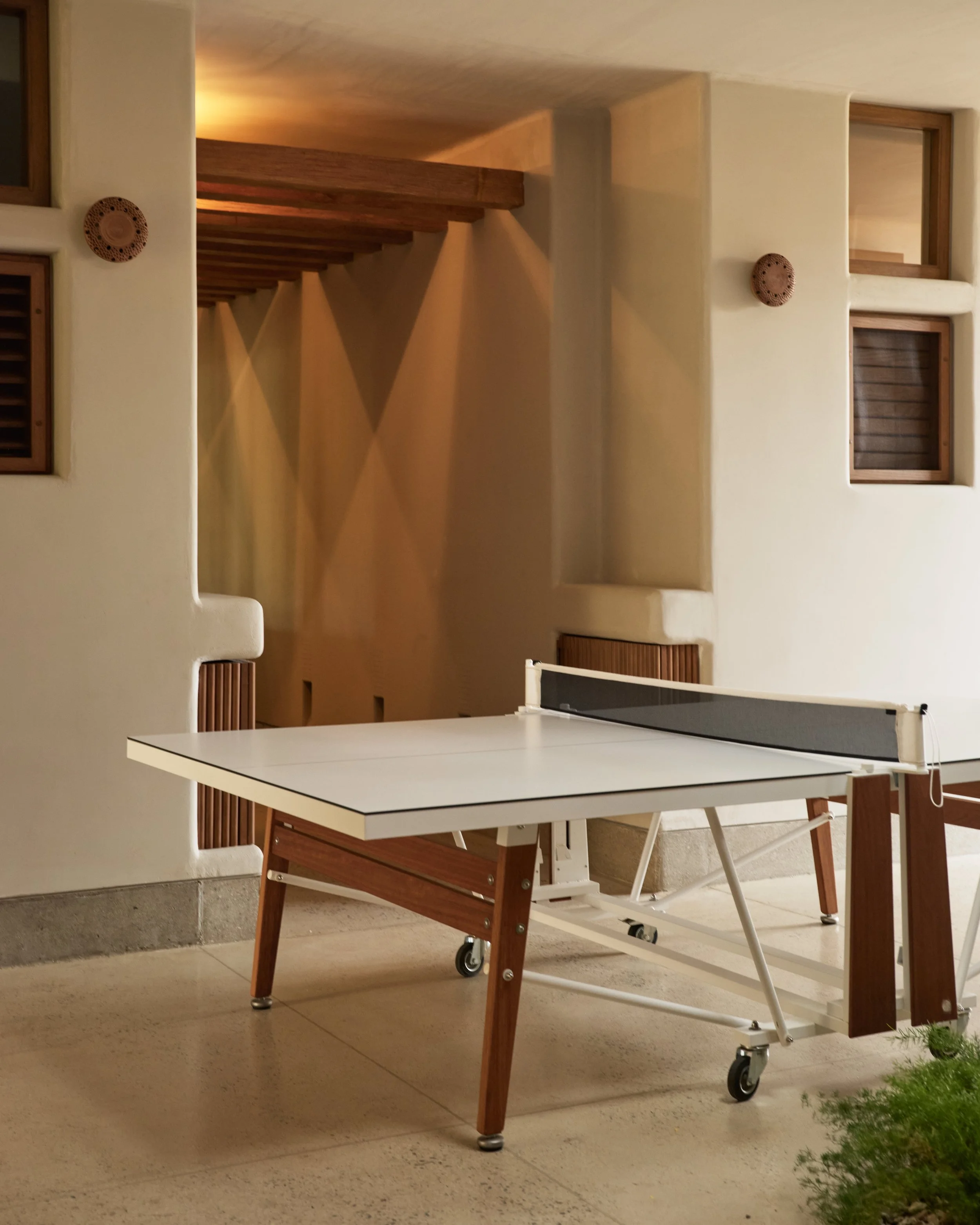 A white ping pong table set up indoors, with a collapsible wooden bench underneath. The room has cream-colored walls, with two small square windows with wooden frames, and decorative wooden plates mounted on the walls. The flooring is light-colored t