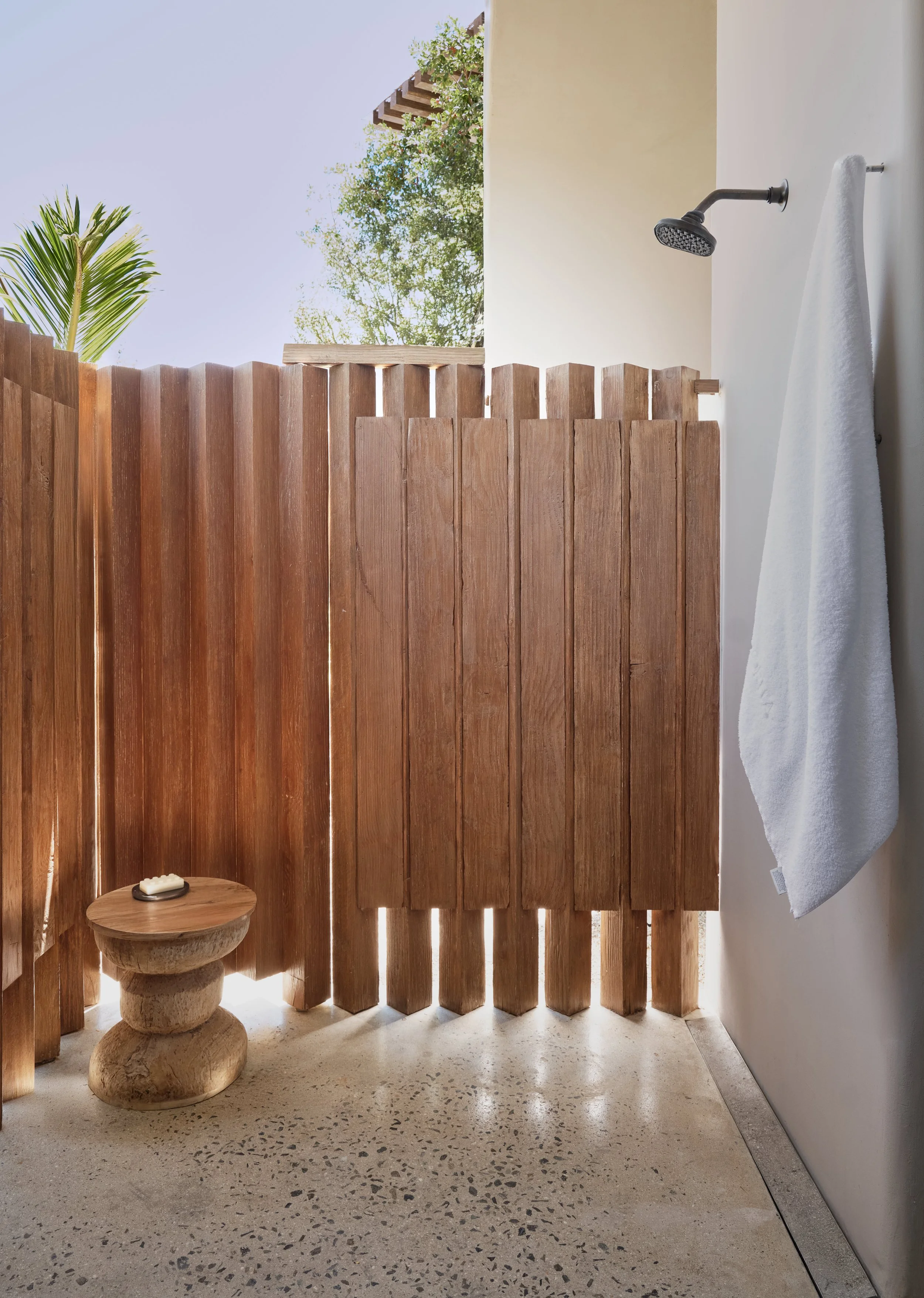 An outdoor shower area with a wooden fence, a hanging towel, a stone side table with soap, and trees visible in the background.