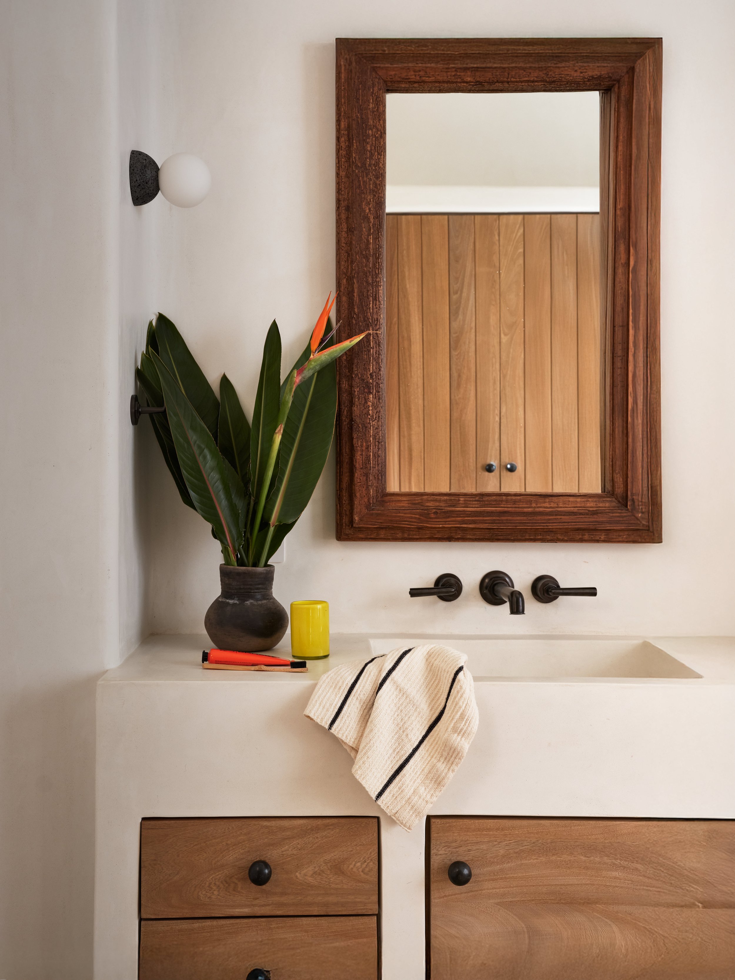 A bathroom sink area with a wooden-framed mirror above a white countertop. On the countertop, there is a potted plant with large green leaves, a yellow cup, and an orange pen. A striped towel is draped over the edge of the sink. The wall has a black 