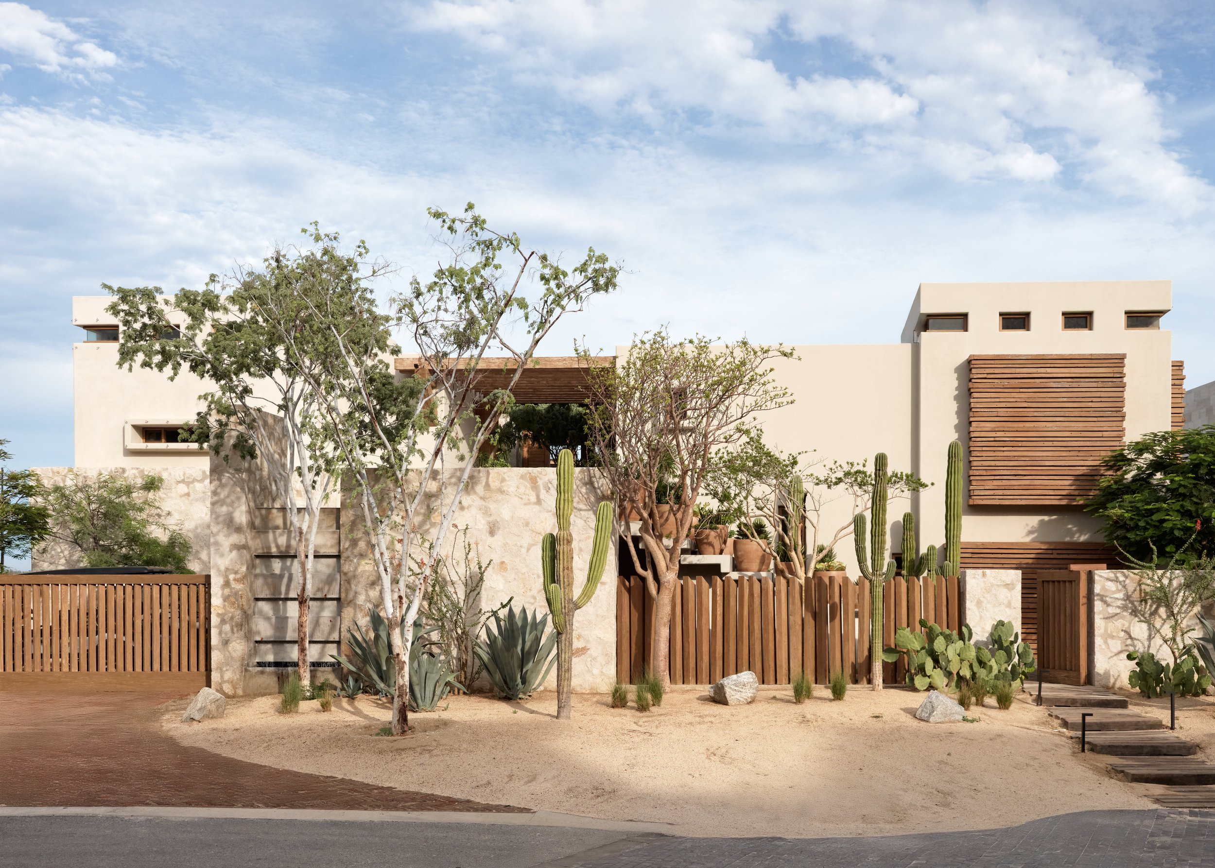 Modern house with desert landscaping, including cacti and drought-tolerant plants, visible behind a wooden fence under a partly cloudy sky.