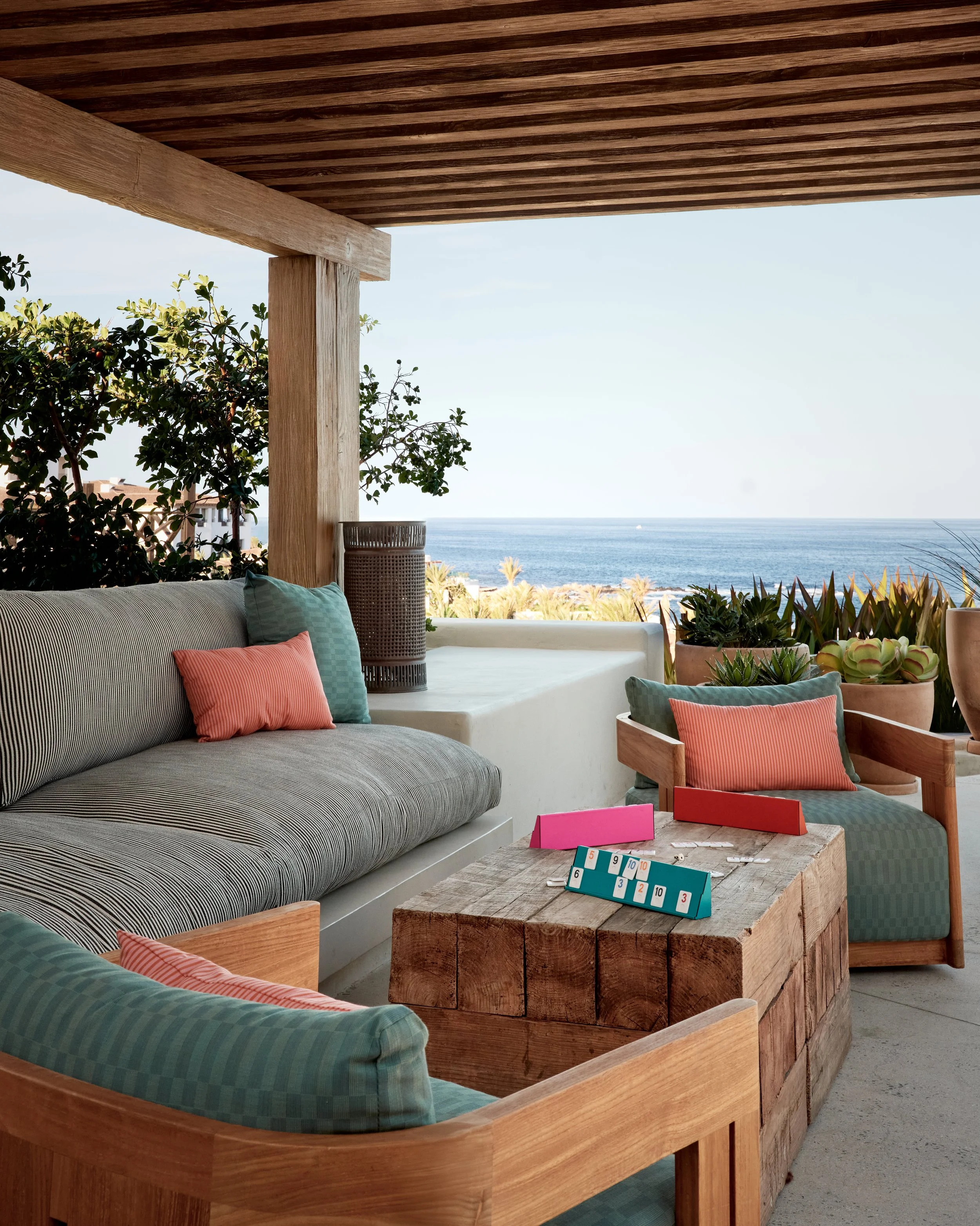 Outdoor patio space with wooden furniture, colorful cushions, potted plants, and a view of the ocean.