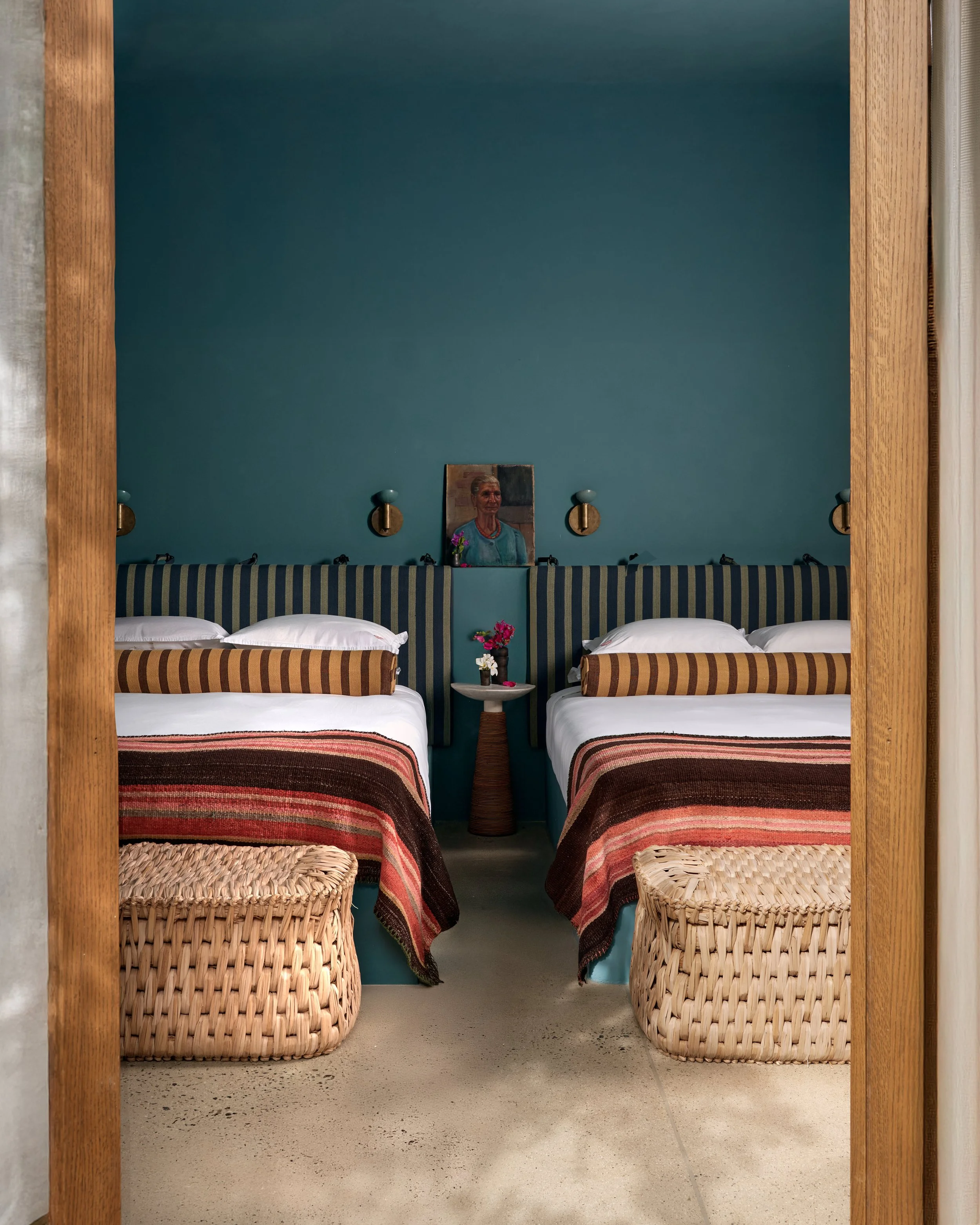 Two beds with striped headboards, blankets, and pillows, set against a teal wall with three wall-mounted lamps, small paintings, and a table with flowers between the beds, viewed from a doorway with wooden frame.