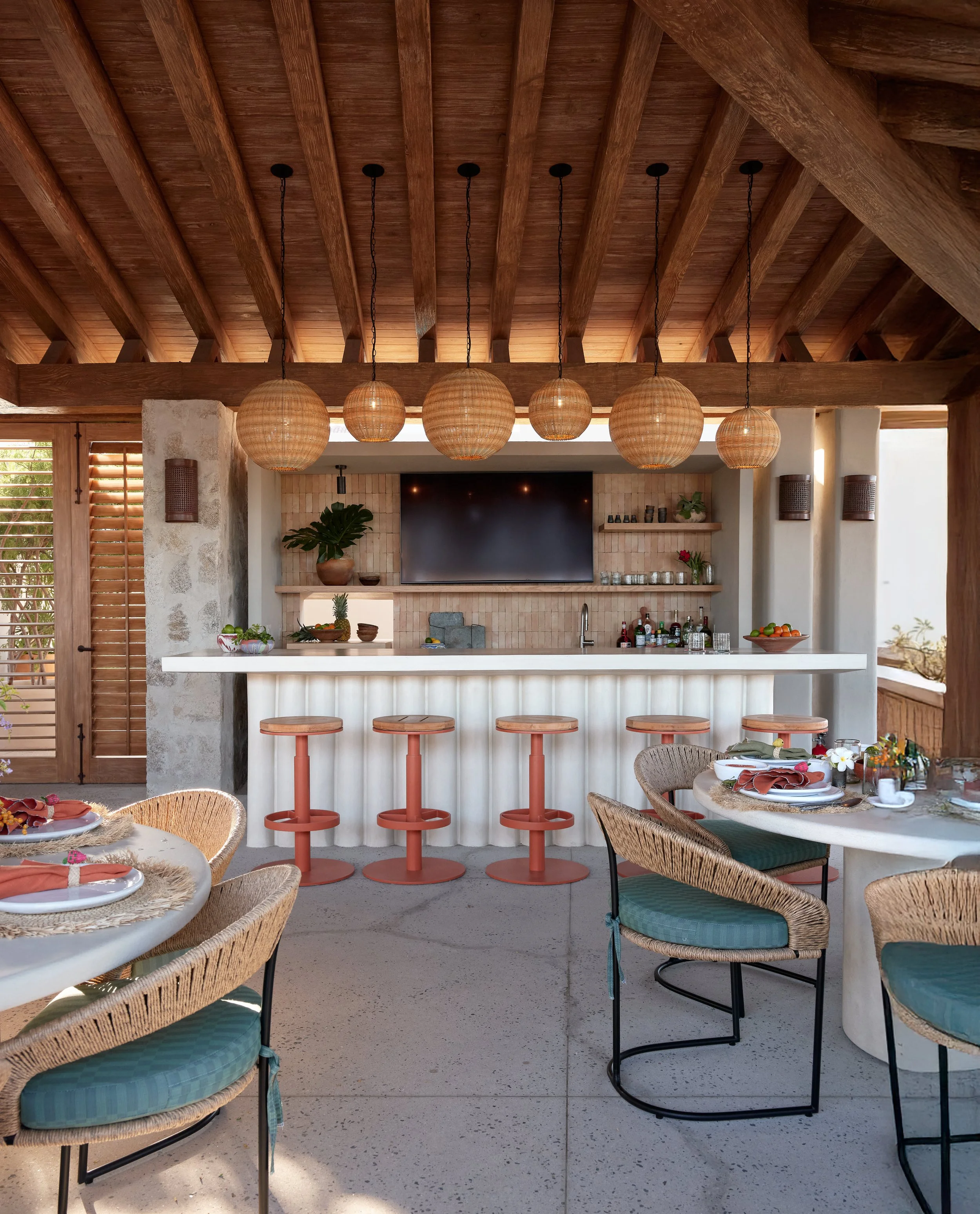 Indoor kitchen with a wooden ceiling, hanging wicker light fixtures, a TV on the wall, a bar with three coral-colored stools, and a dining table with wicker chairs set for a meal.