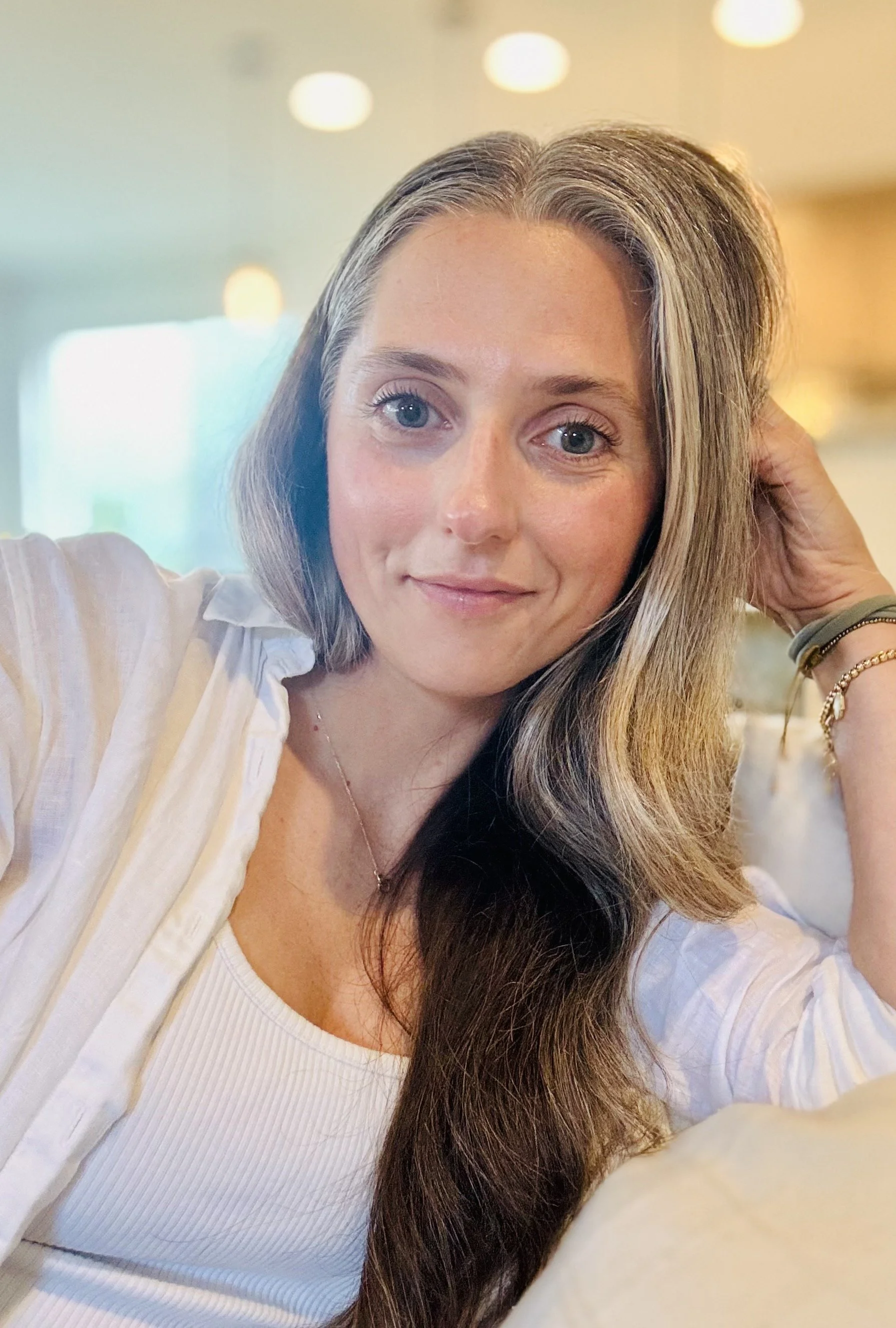 Close-up portrait of a smiling woman with long brown hair wearing a white blouse and bracelets