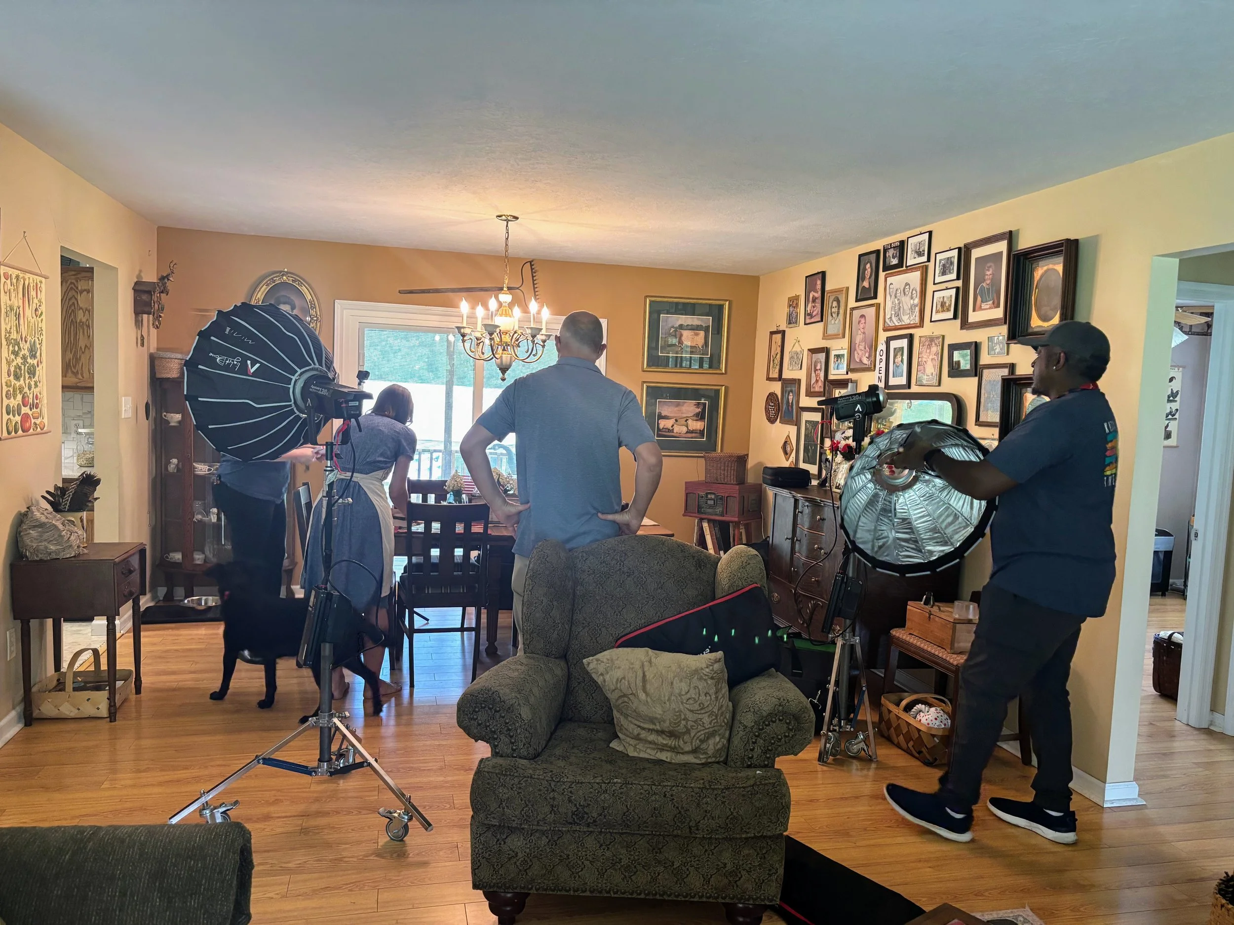A family or group of people with a dog in a dining and living room, with film or photography equipment set up, and walls decorated with framed photos and art.
