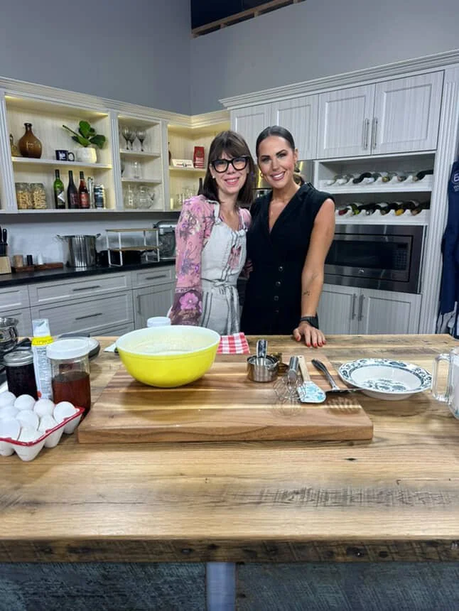 Two women standing in a kitchen behind a wooden countertop with baking ingredients and utensils, smiling at the camera.